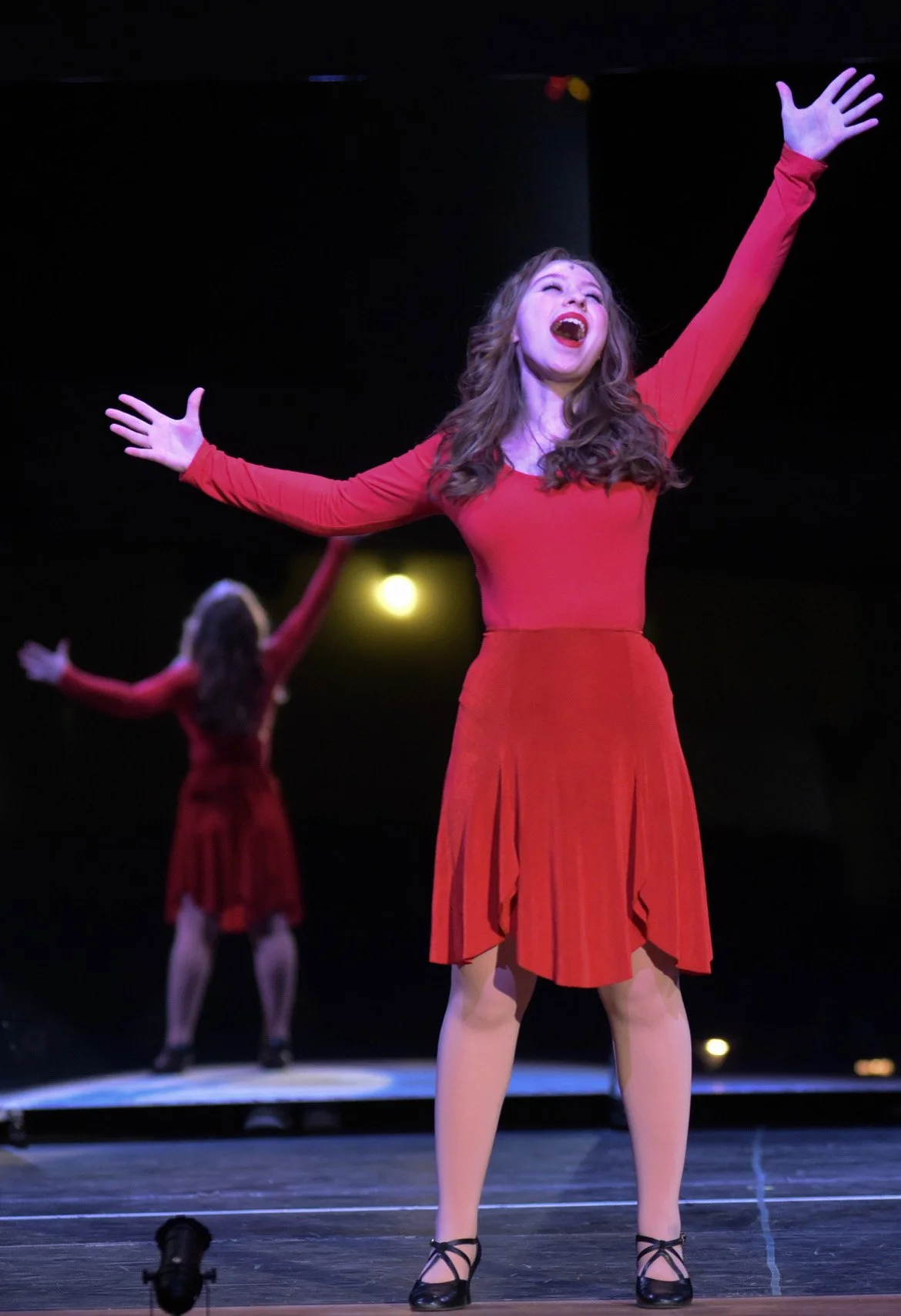 Cassie in A chorus line. A woman in a red dress singing and dancing on stage with arms outstretched, with another woman performing in the background.