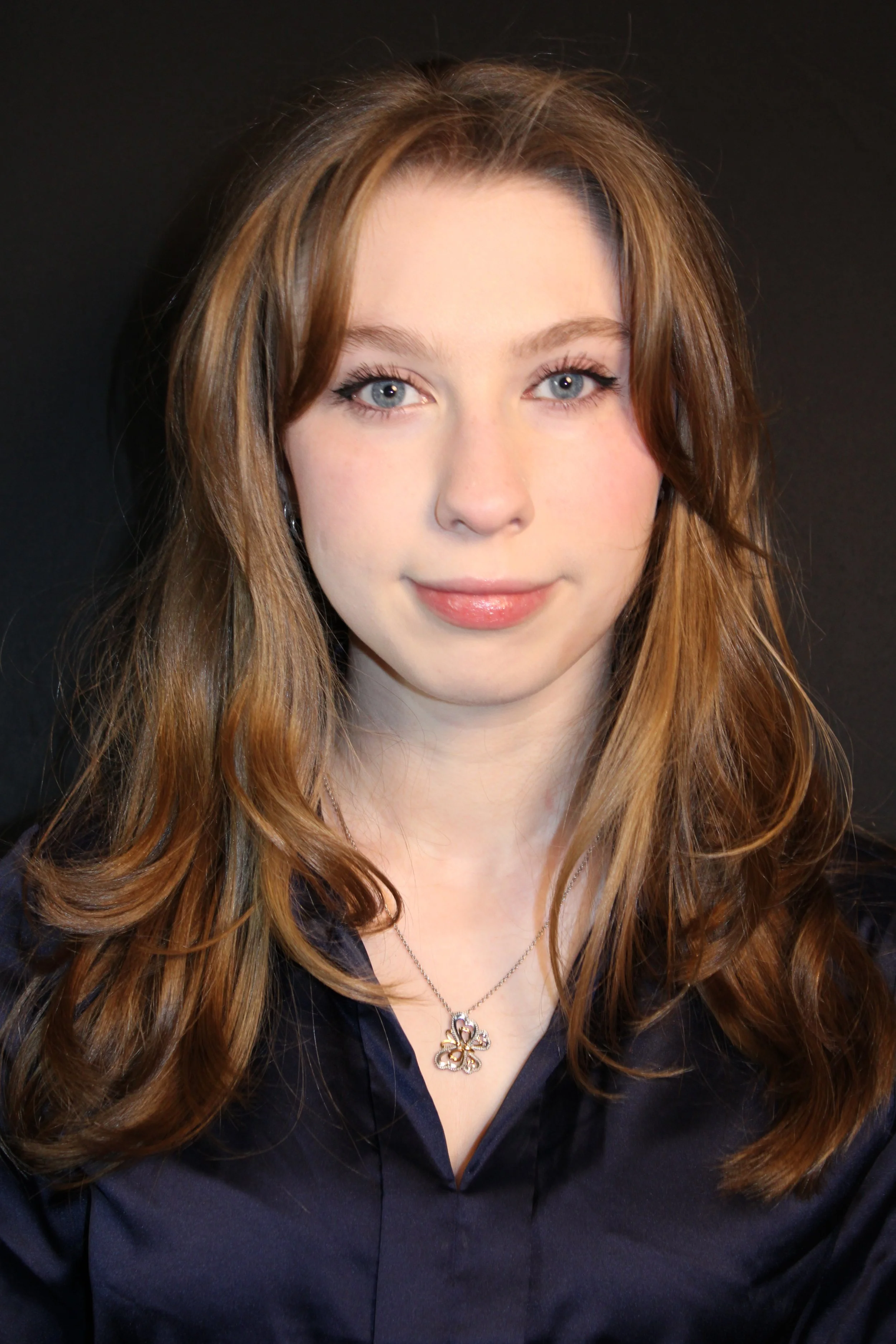 Gorgeous beautiful girl next door or bombshell versatile appearance. Can play childlike, teens, or twenties. A young woman with long, wavy, light brown hair and blue eyes, smiling softly against a black background.