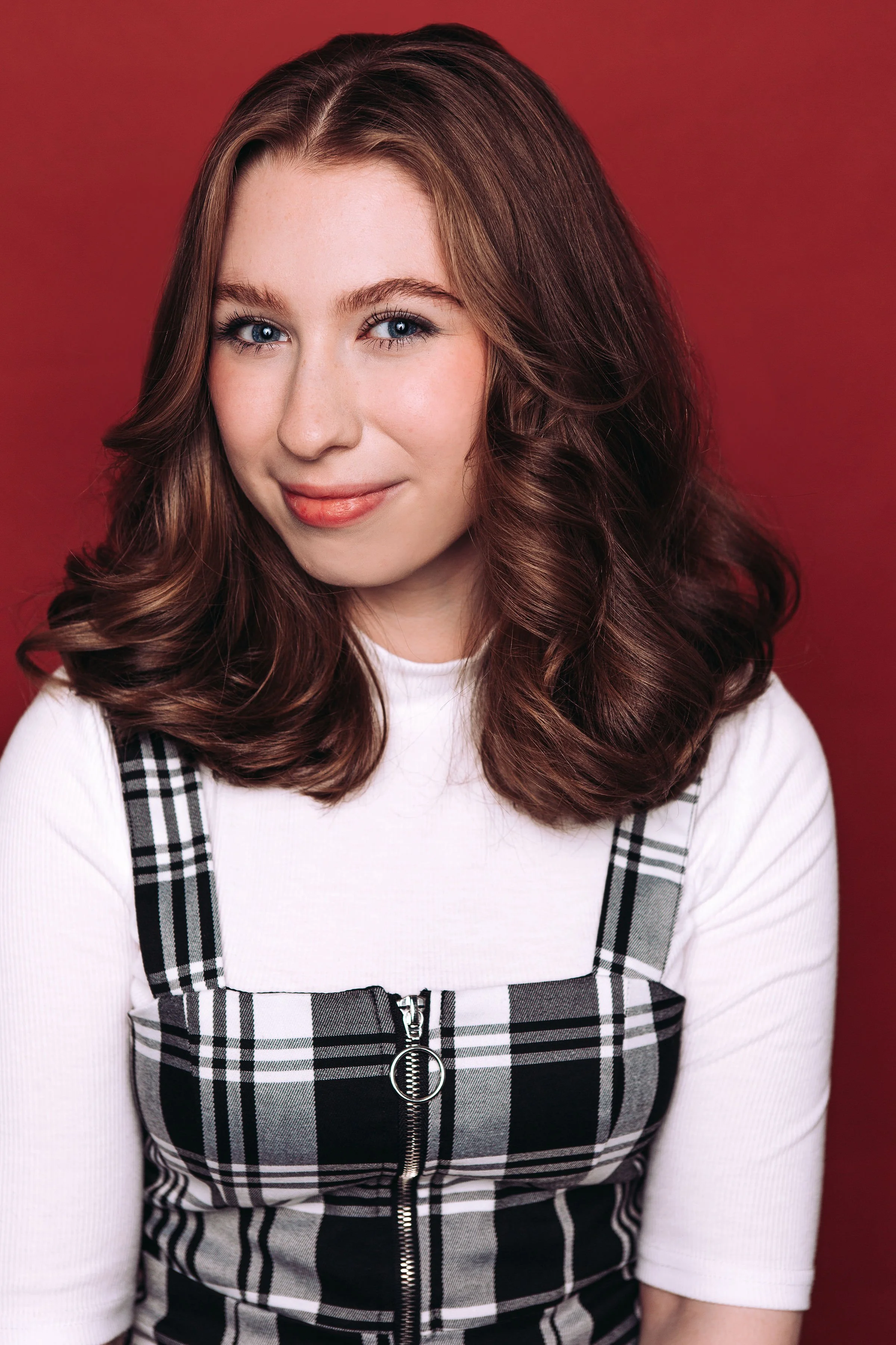 Grace is an age versatile young woman with shoulder-length brown hair, blue eyes, and light skin, smiling slightly, wearing a white top and a plaid black and white dress with a zipper, against a red background.