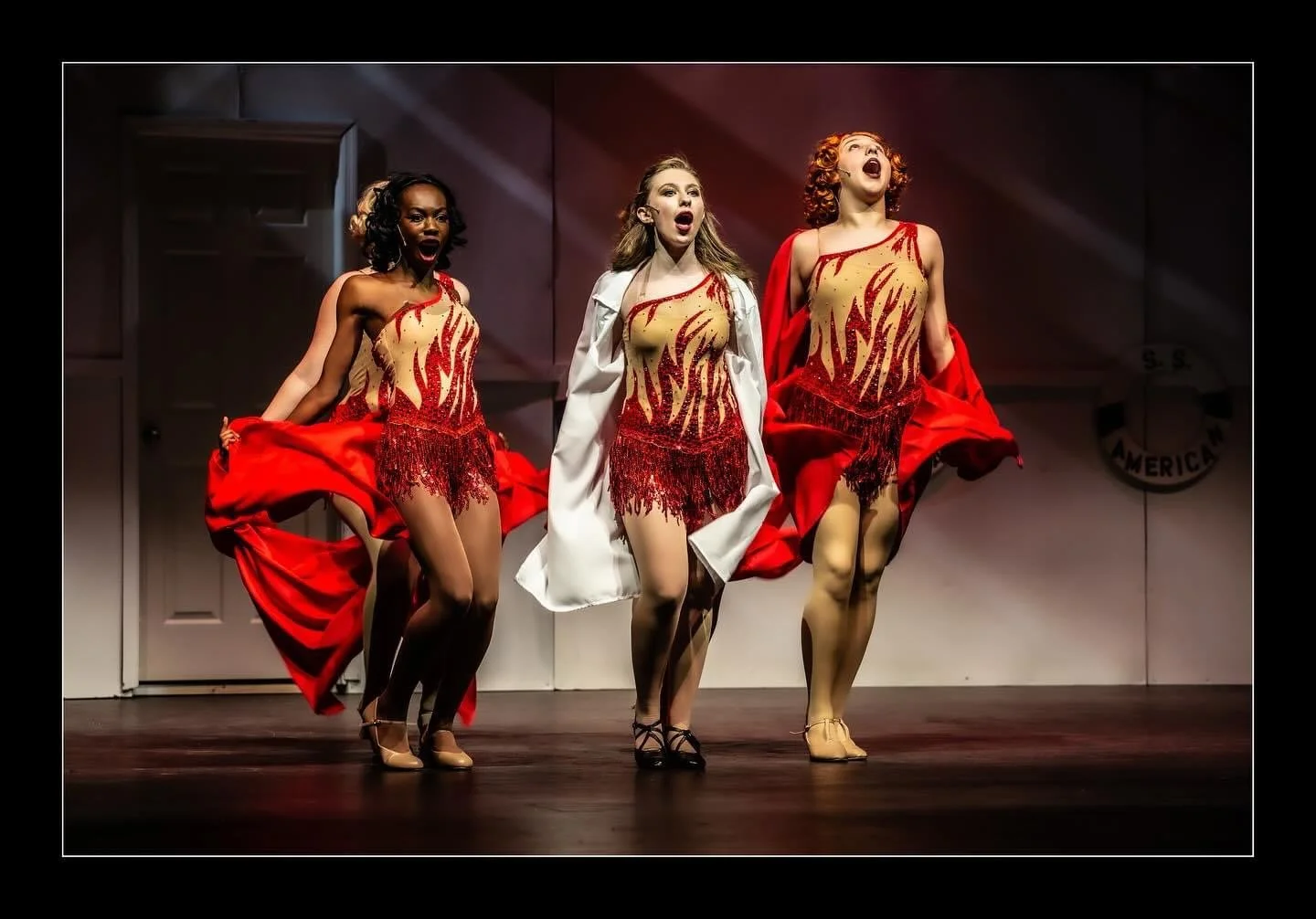 Reno Sweeney performance of Blow Gabriel Blow in Anything Goes. Three women performing on stage in red and gold costumes, singing or dancing with red jackets draped over their shoulders.