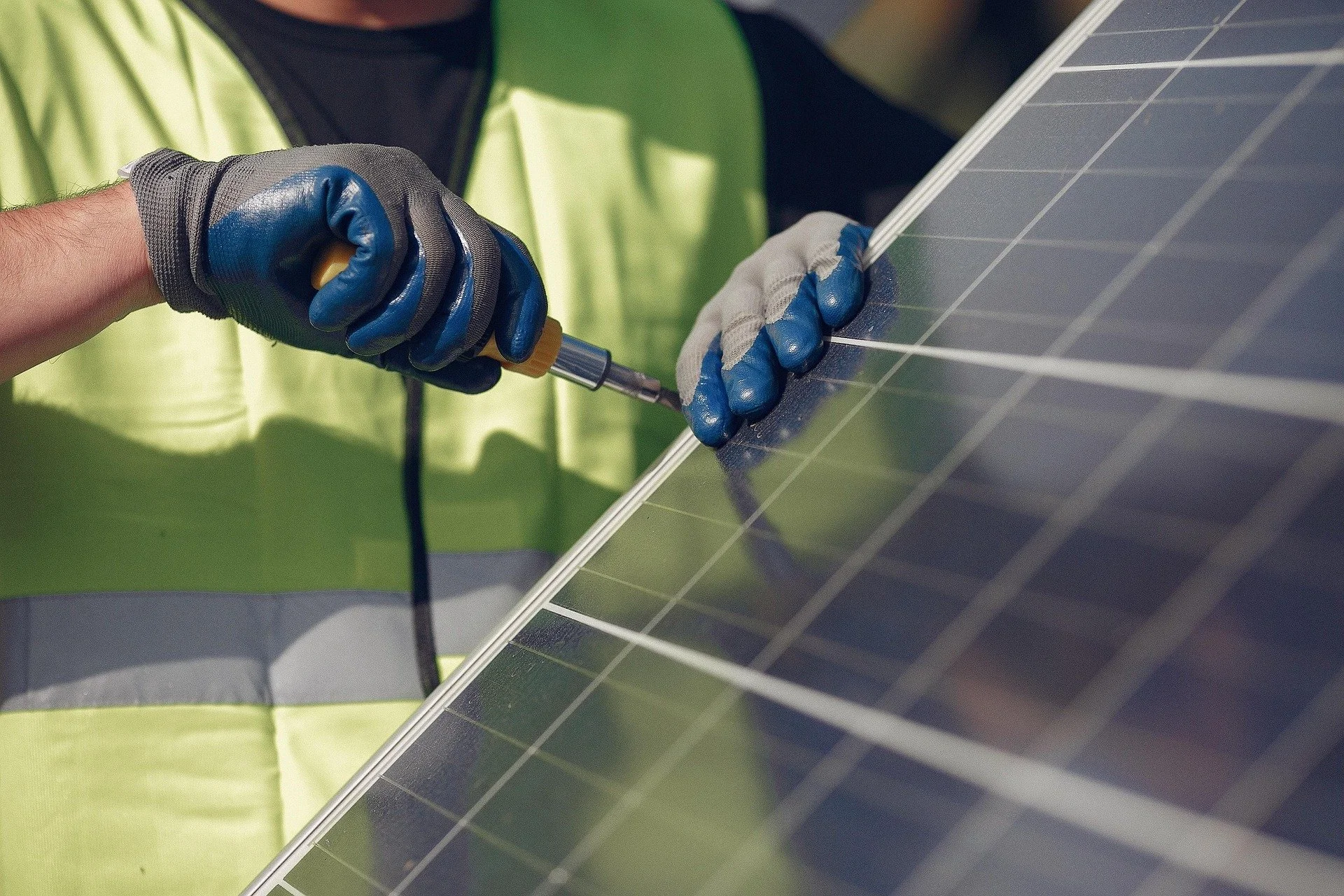 A person wearing gray and blue gloves screws into a solar panel with a screwdriver.