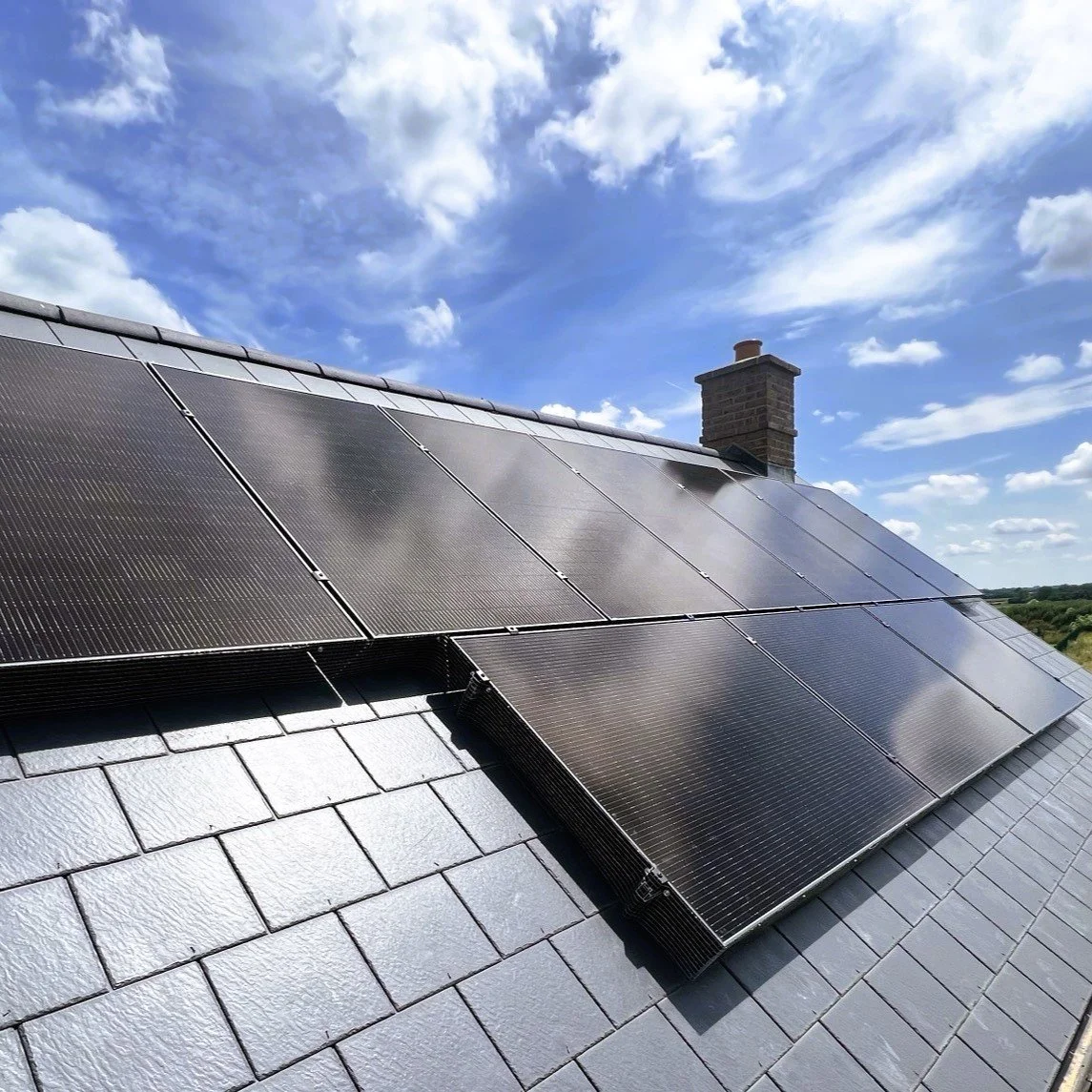 Black solar panels installed on a gray shingle roof under a blue sky with scattered clouds and a brick chimney in the background.
