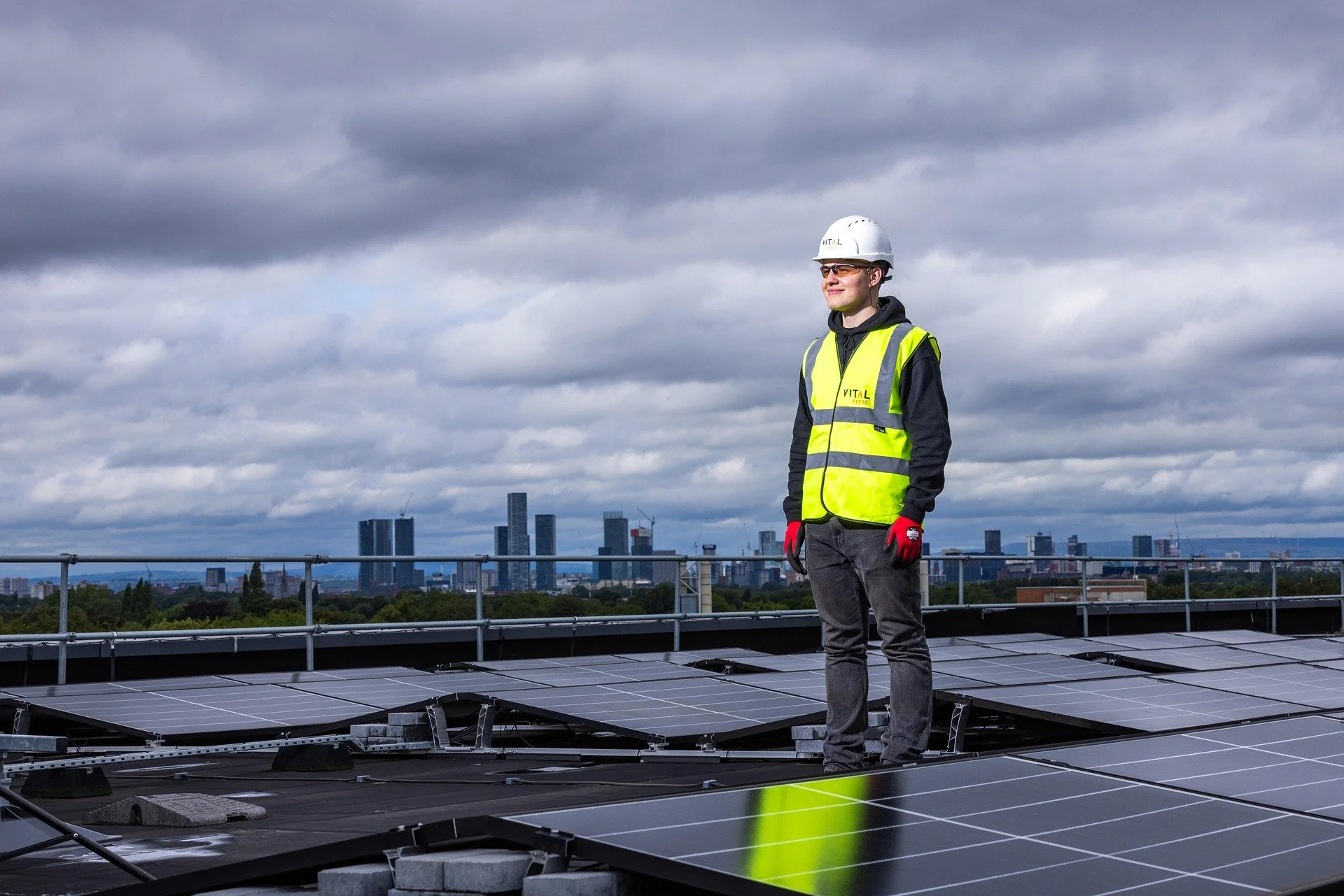 A person in safety gear standing on a rooftop with solar panels, city skyline in the background, cloudy sky above.