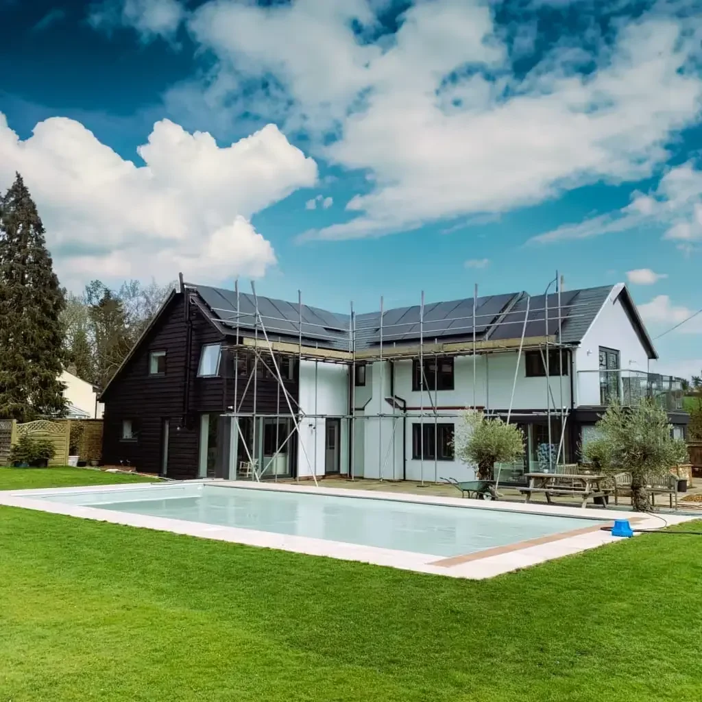 A two-story house under renovation with scaffolding around it, a swimming pool in the backyard, and solar panels on the roof, with a grassy lawn and cloudy sky