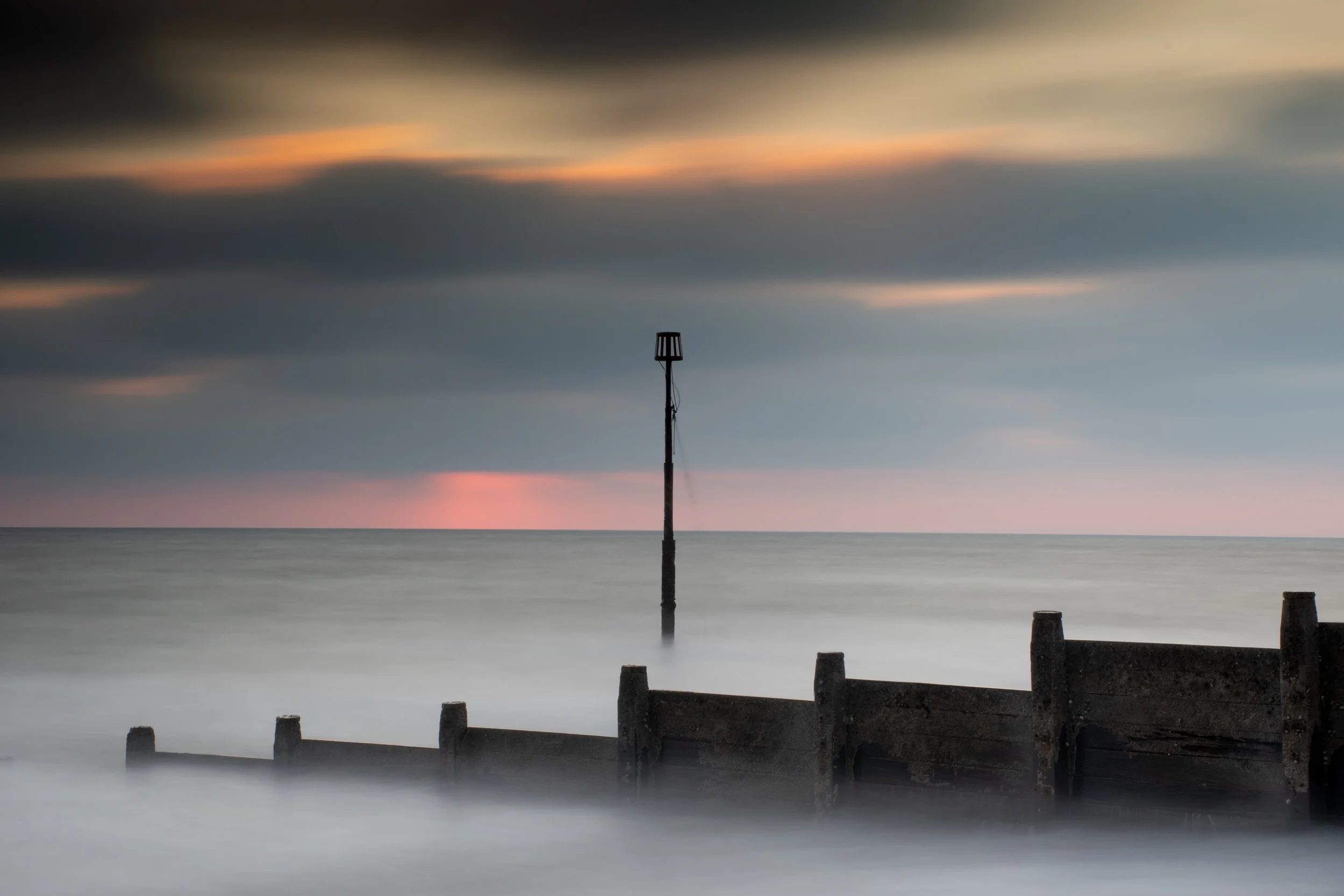 A sunset over the ocean with a cloudy sky, a tall navigation marker in the water, and a wooden seawall in the foreground.