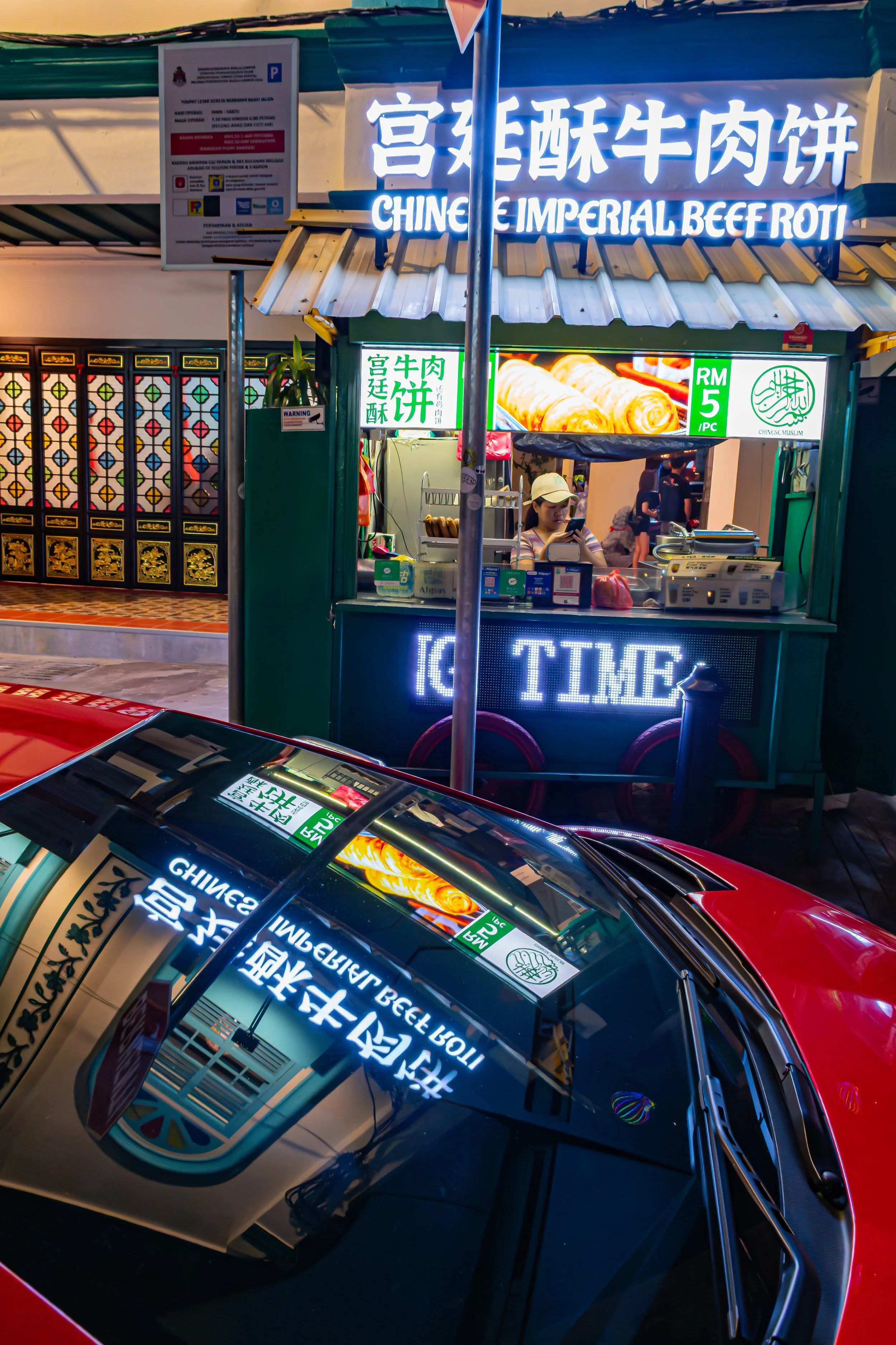 A street food stall with a neon sign that reads 'Chine Imperial Beef Roti' and Chinese characters, offering beef roti for RM5 each. The stall is attended by a woman wearing a cap, and there are people in the background. Reflected in the car's windshield is the stall's sign and a section of a colorful wall.