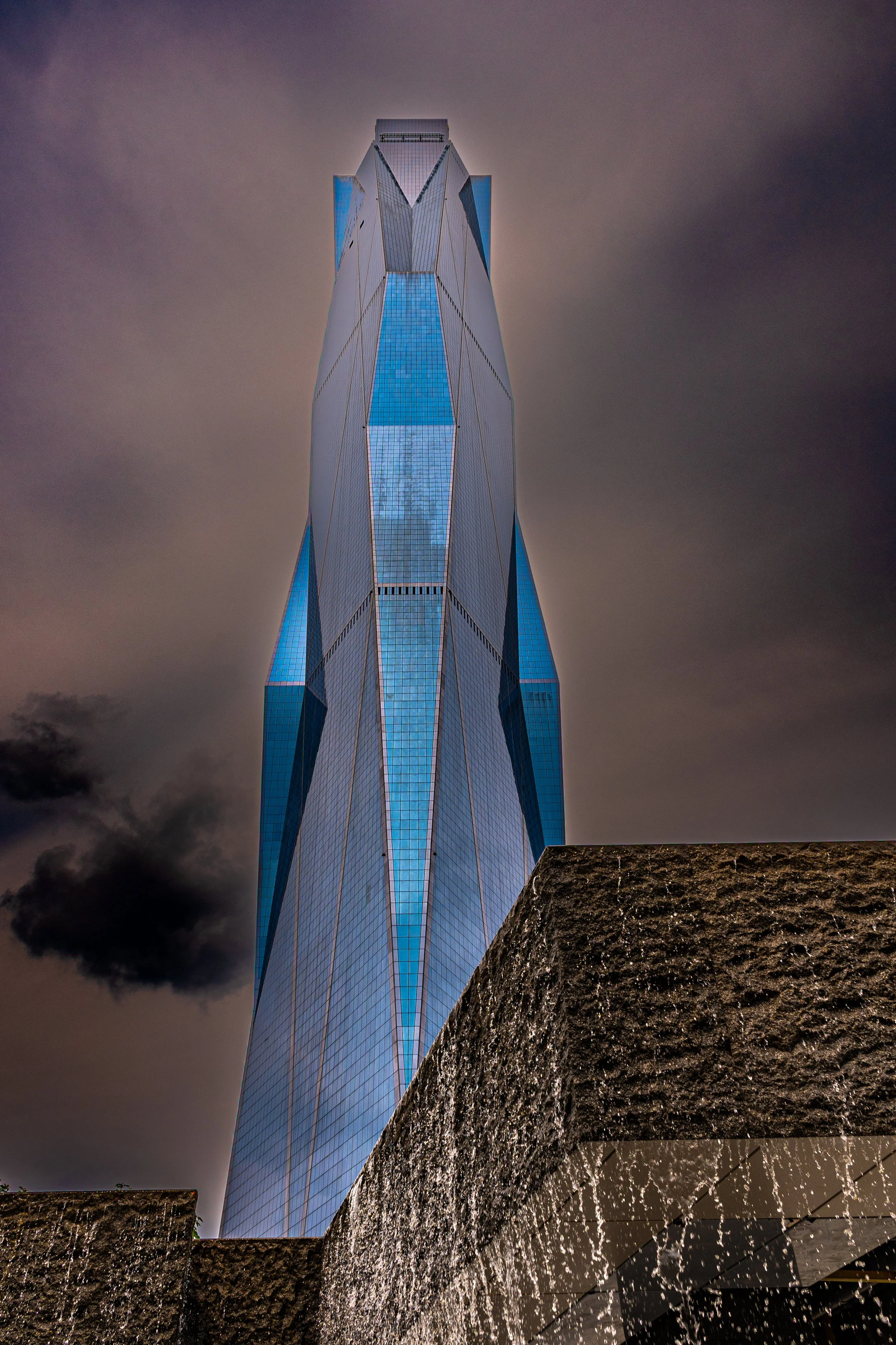 A tall, modern skyscraper with a geometric design and reflective glass exterior, seen against a dark, cloudy sky, with a stone water fountain in the foreground.
