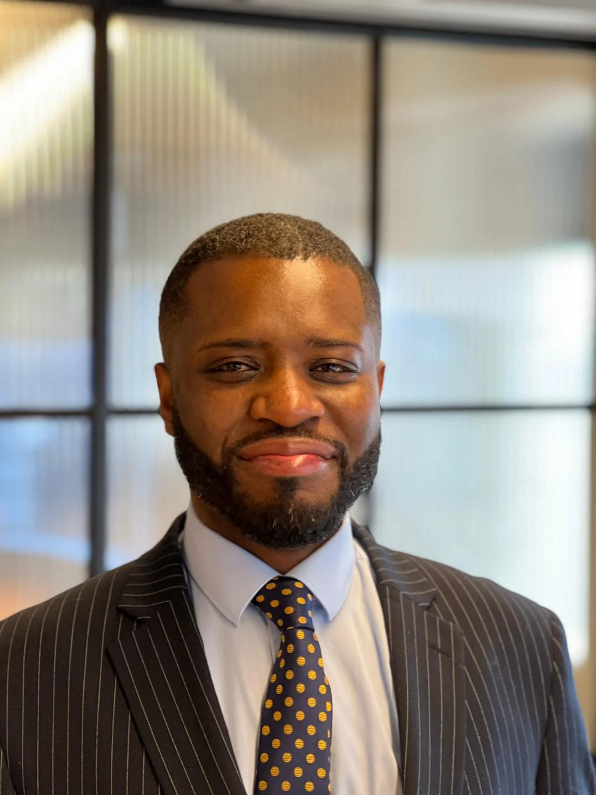 Portrait of a man wearing a dark pinstripe suit, white shirt, and a navy tie with yellow polka dots, standing indoors with a glass wall in the background.