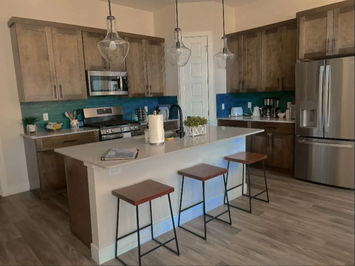 Modern kitchen with wooden cabinets, stainless steel appliances, a white island with three stools, pendant lights, and a teal backsplash.