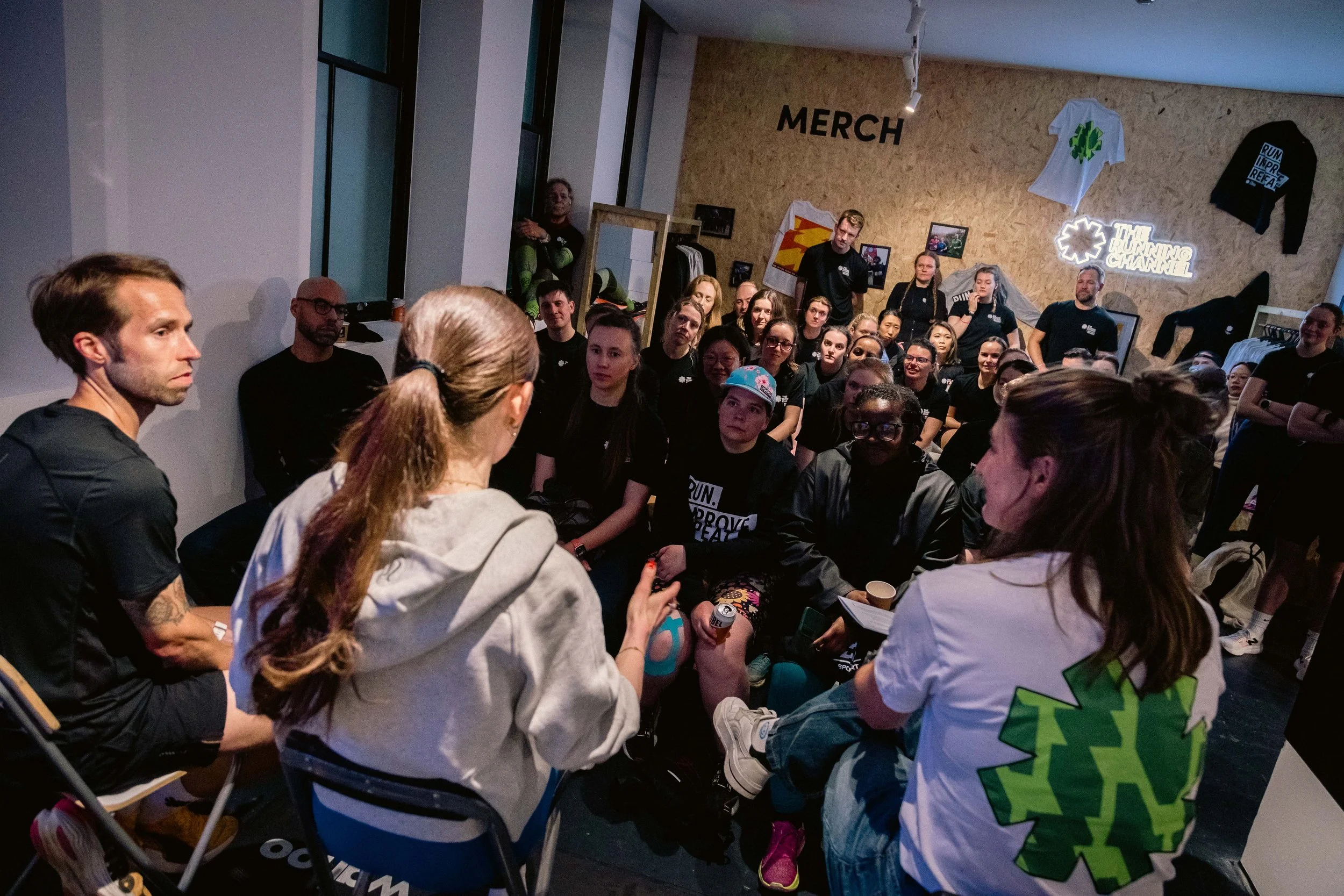 Group of people attending a presentation in a modern, decorated room with merchandise on the wall, including T-shirts and signs, and a large window on the left side.
