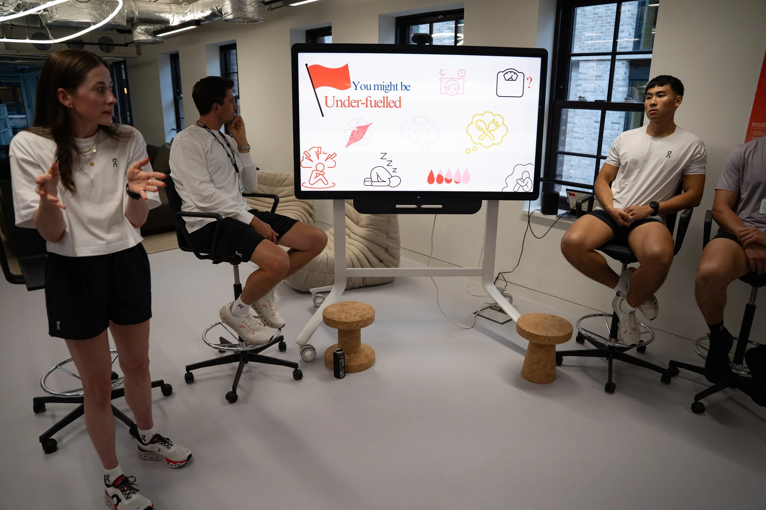 A group of fitness trainers in a studio, part of a presentation on signs of dehydration, with one woman speaking and three men seated on adjustable stools, a digital display behind them showing related illustrations and text.