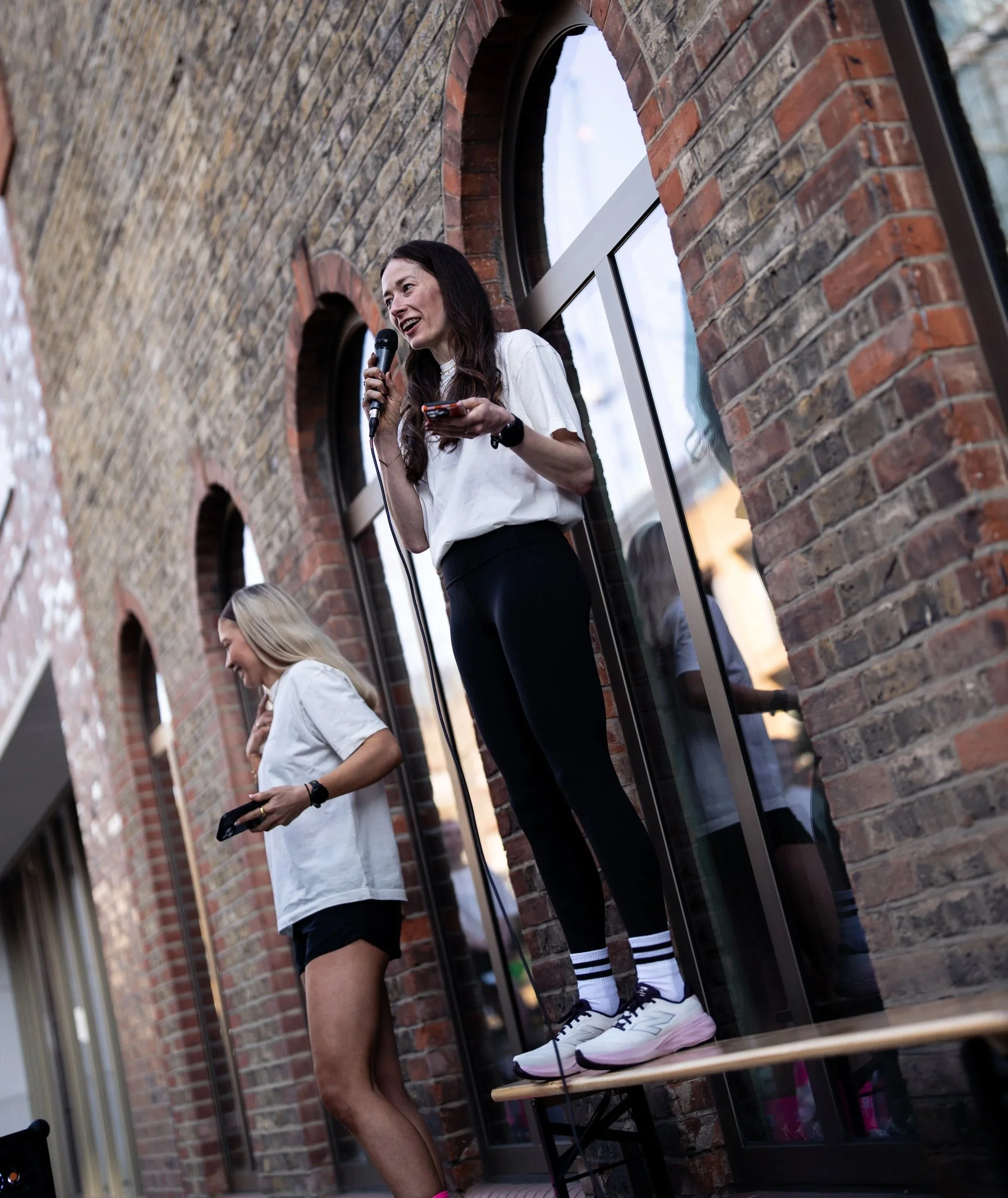 A woman speaking into a microphone while standing on a small platform outdoors next to a brick building, with another woman standing nearby holding a remote control.