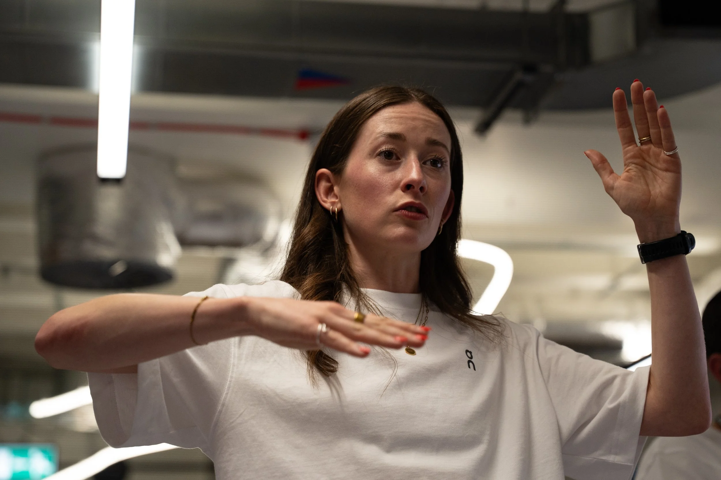 A woman with long brown hair wearing a white t-shirt, rings, and a watch raises her hands while speaking or explaining in an indoor setting with ceiling lights and industrial ceiling elements.