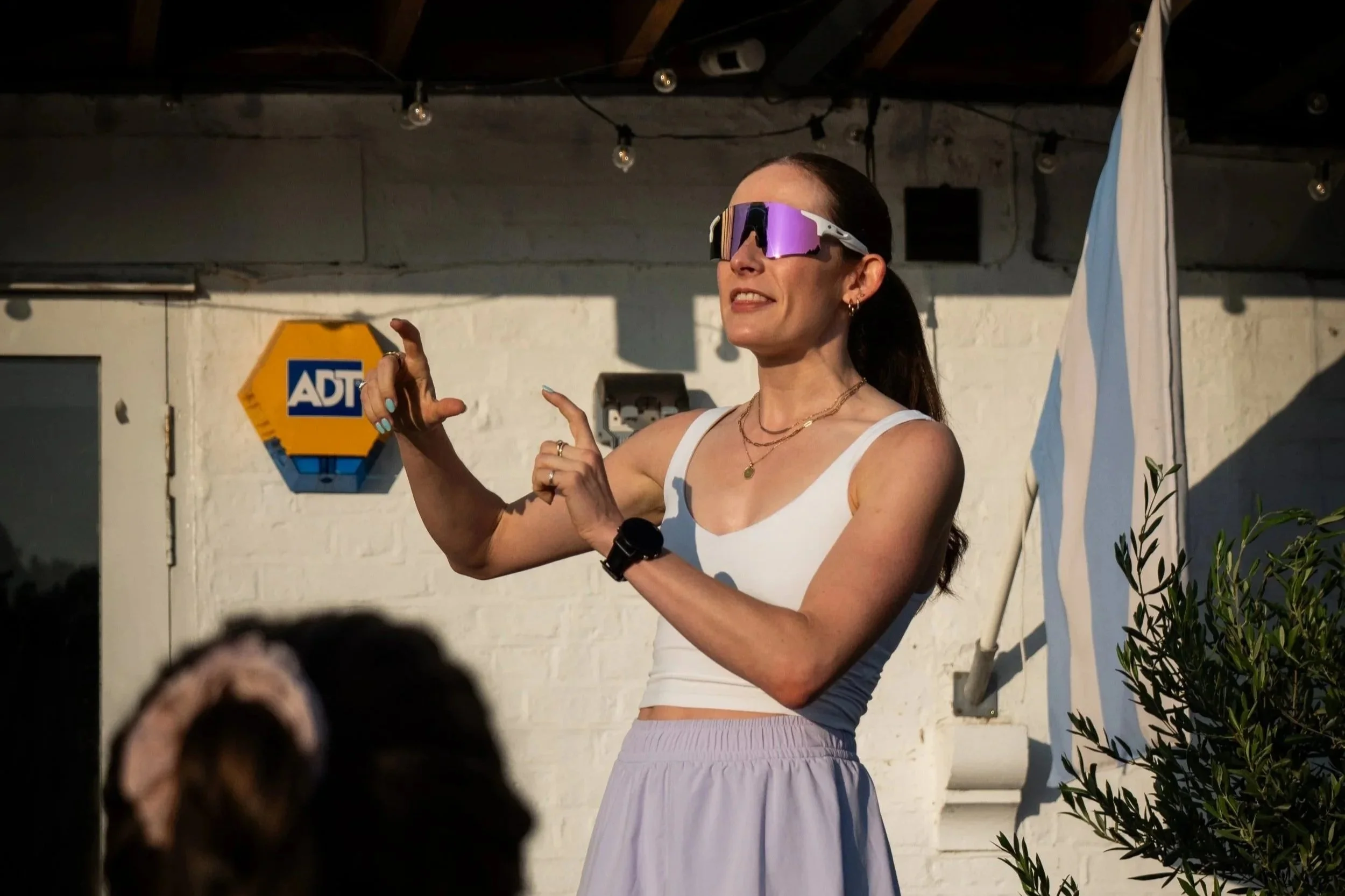 A woman wearing sunglasses, a white tank top, and a light pink skirt, appears to be speaking or presenting outdoors. She has her hair tied back, and is wearing jewelry and a smartwatch. In the background, there's a white brick wall with a yellow and blue ADT security sign and a standing flag.
