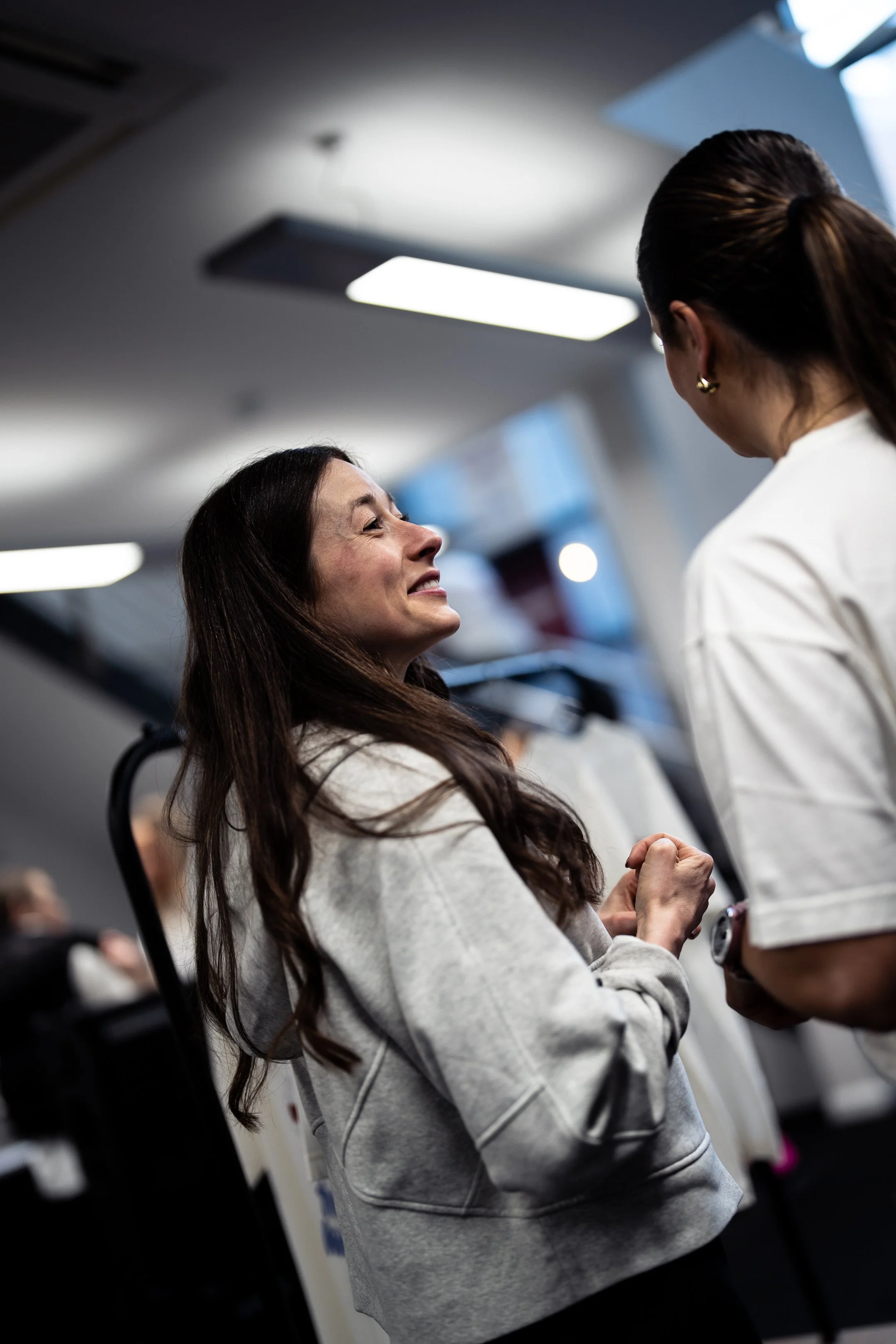 A woman with long brown hair smiling and clenching her fists in excitement while talking to another woman in an airport or train station.