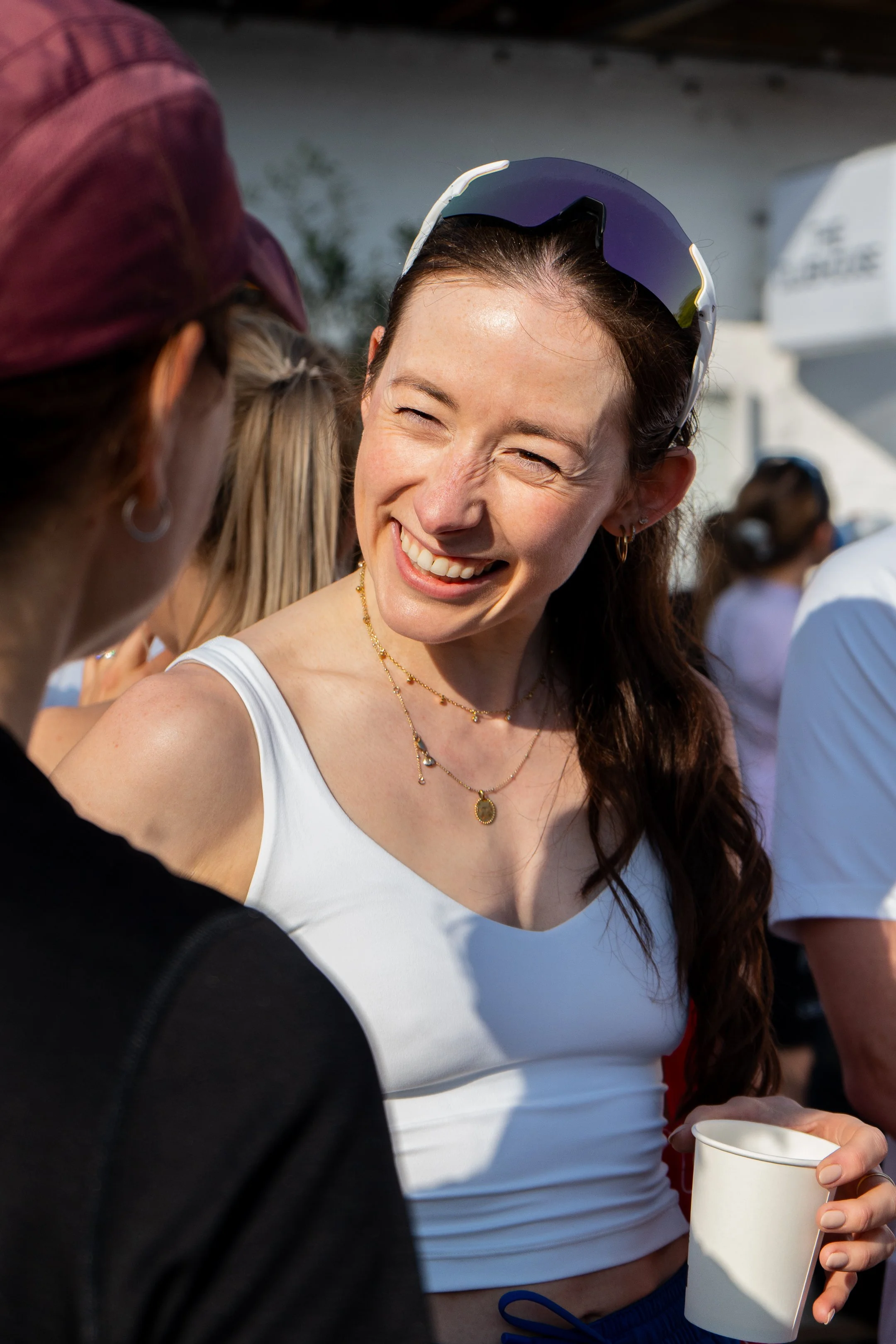 A woman smiling and laughing outdoors, holding a white disposable cup, wearing a white tank top, sunglasses on her head, and jewelry, surrounded by other people.