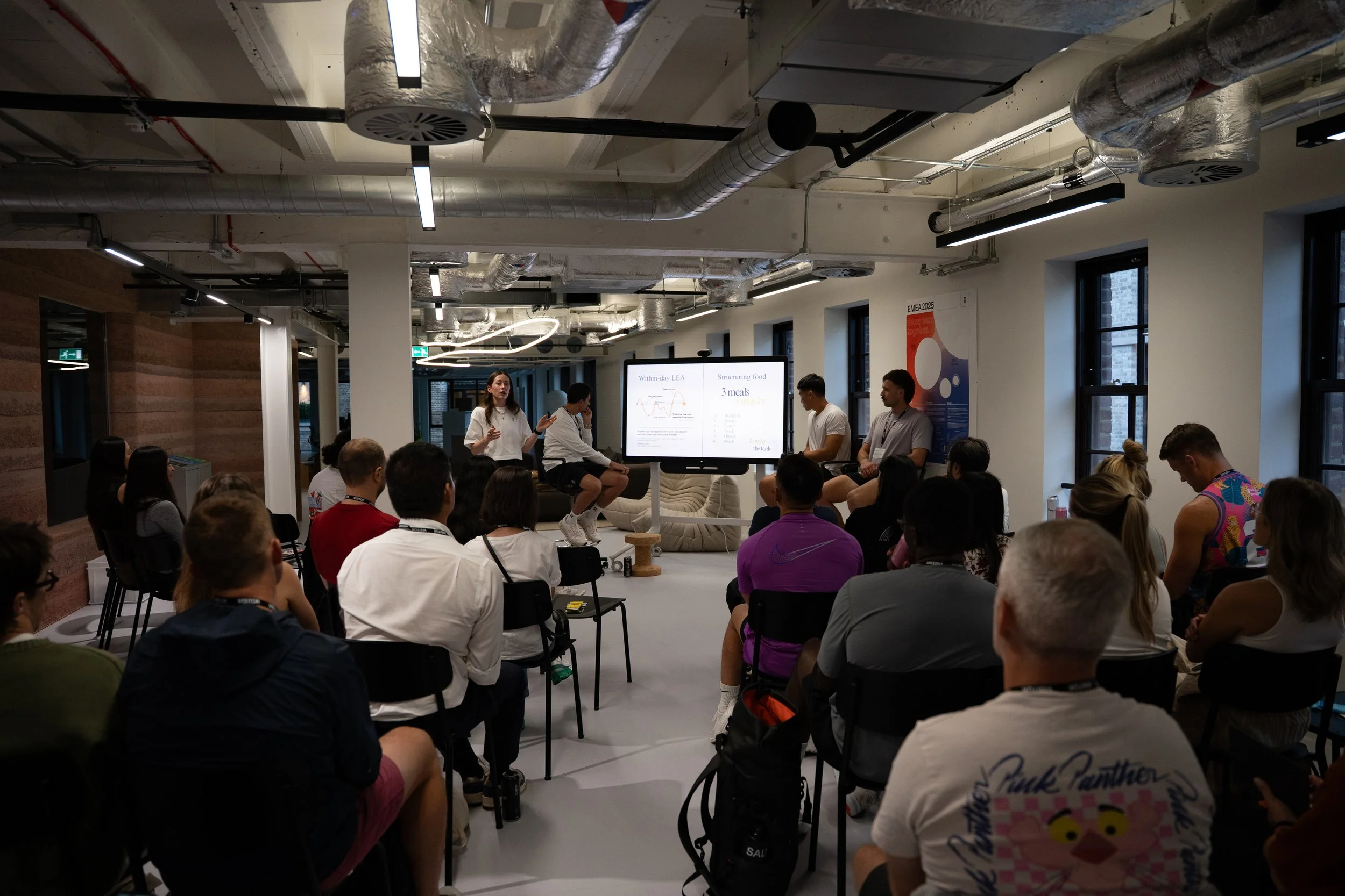A group of people seated in chairs watching a panel discussion in a modern conference room with exposed ductwork ceiling, large windows, and a screen displaying a presentation.