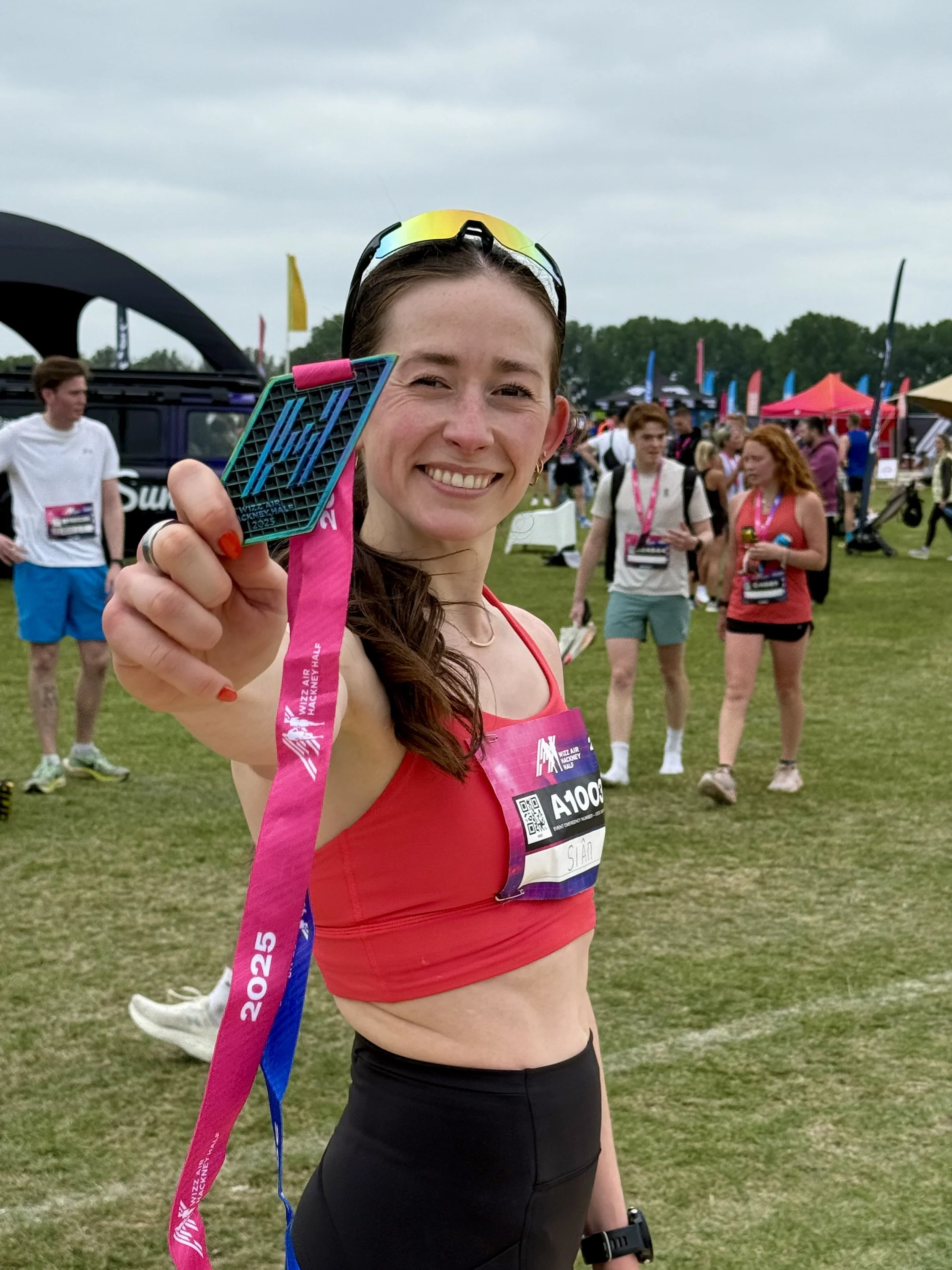 A young woman in a red sports top and black leggings holds up a finisher medal at a marathon event, smiling, with other runners and event tents in the background.