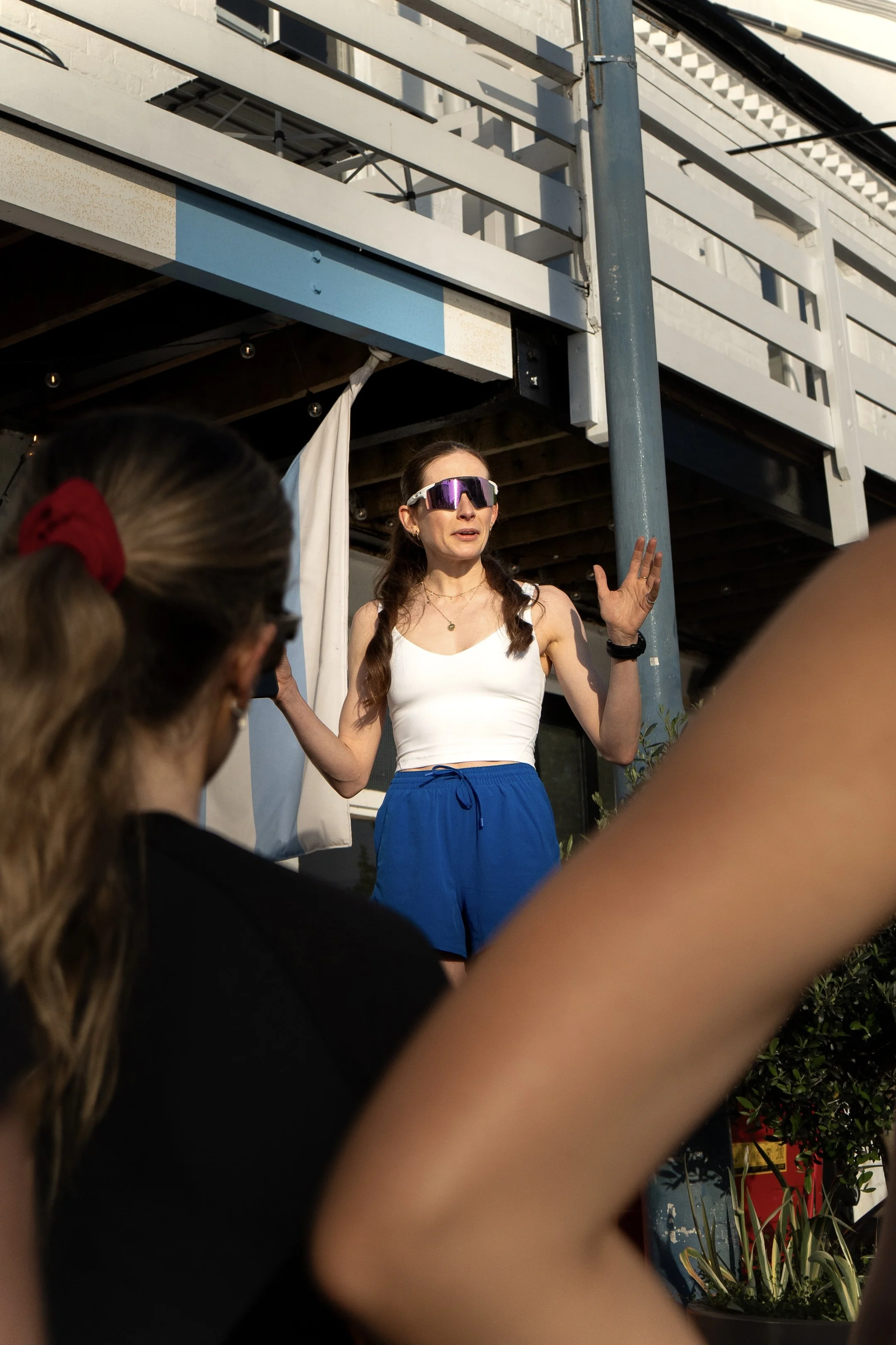 A woman with long brown hair, wearing sunglasses, a white sleeveless top, and blue shorts, speaking to an audience outdoors. Multiple people in the foreground are listening to her.