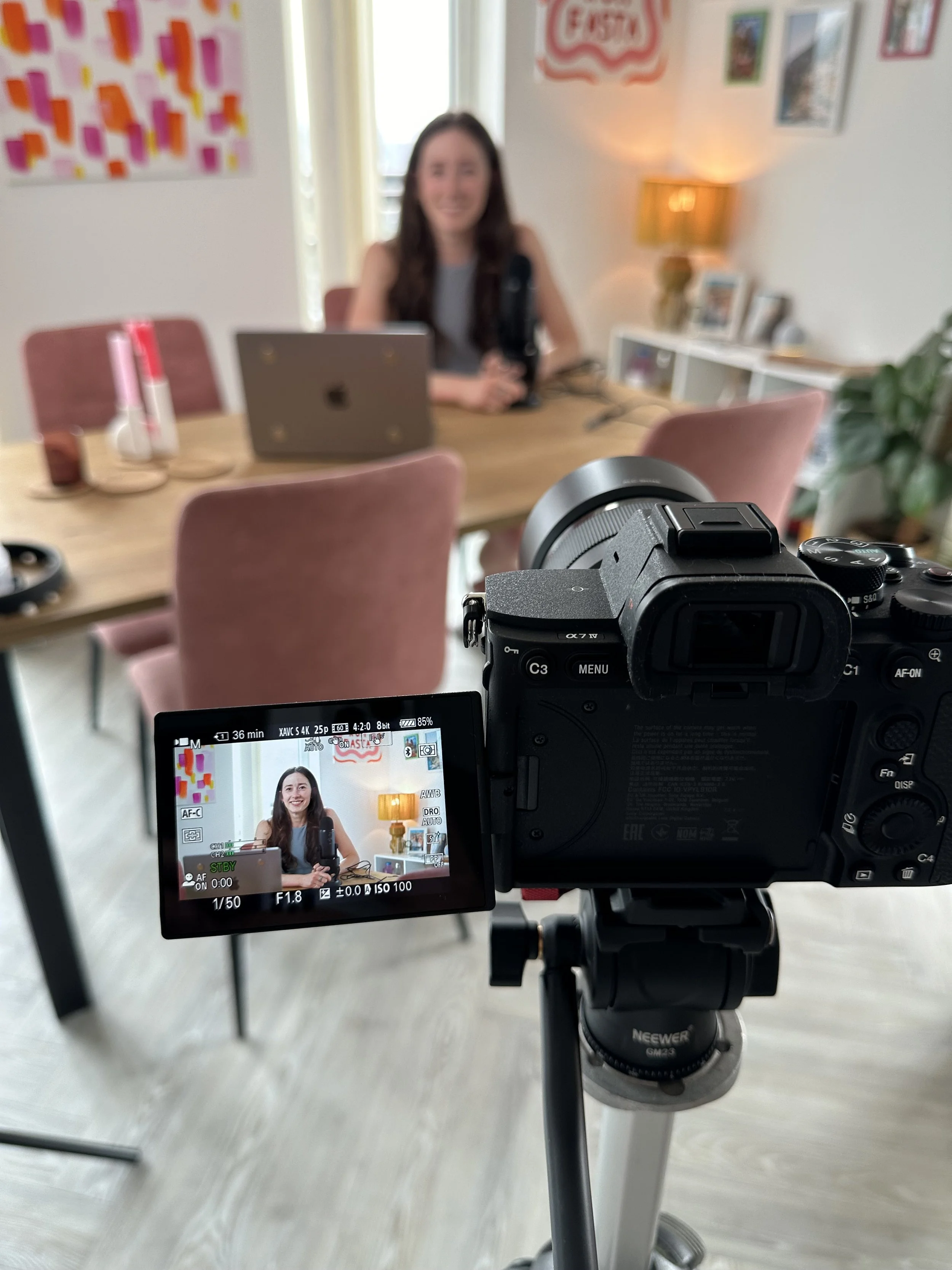 A camera on a tripod capturing a woman sitting at a dining table with a laptop in front of her, in a bright room with artwork and decorations on the walls.