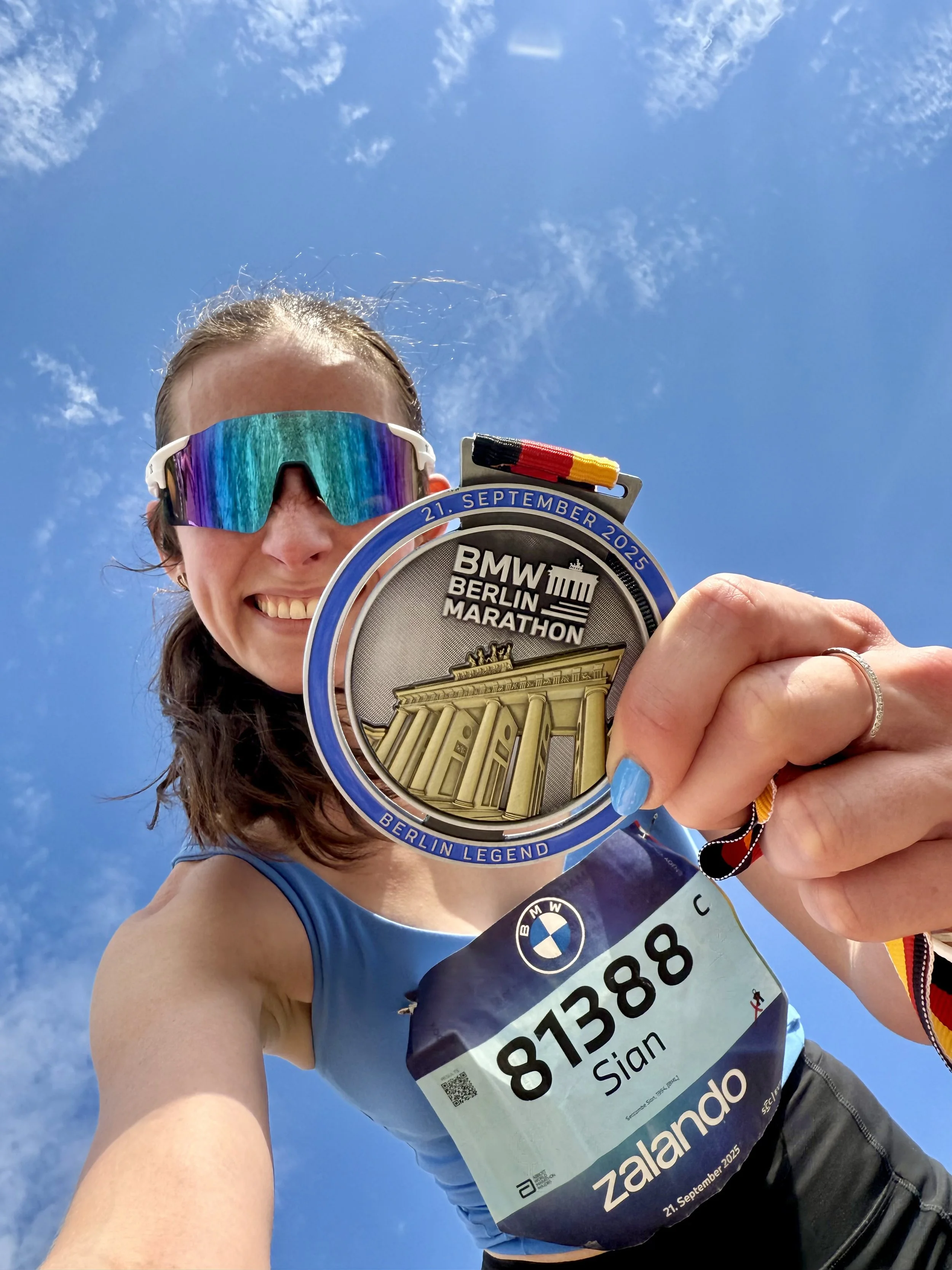 A woman wearing sunglasses and a blue athletic top holding up a finisher's medal from the BMW Berlin Marathon with a race bib. The sky is clear with few clouds.