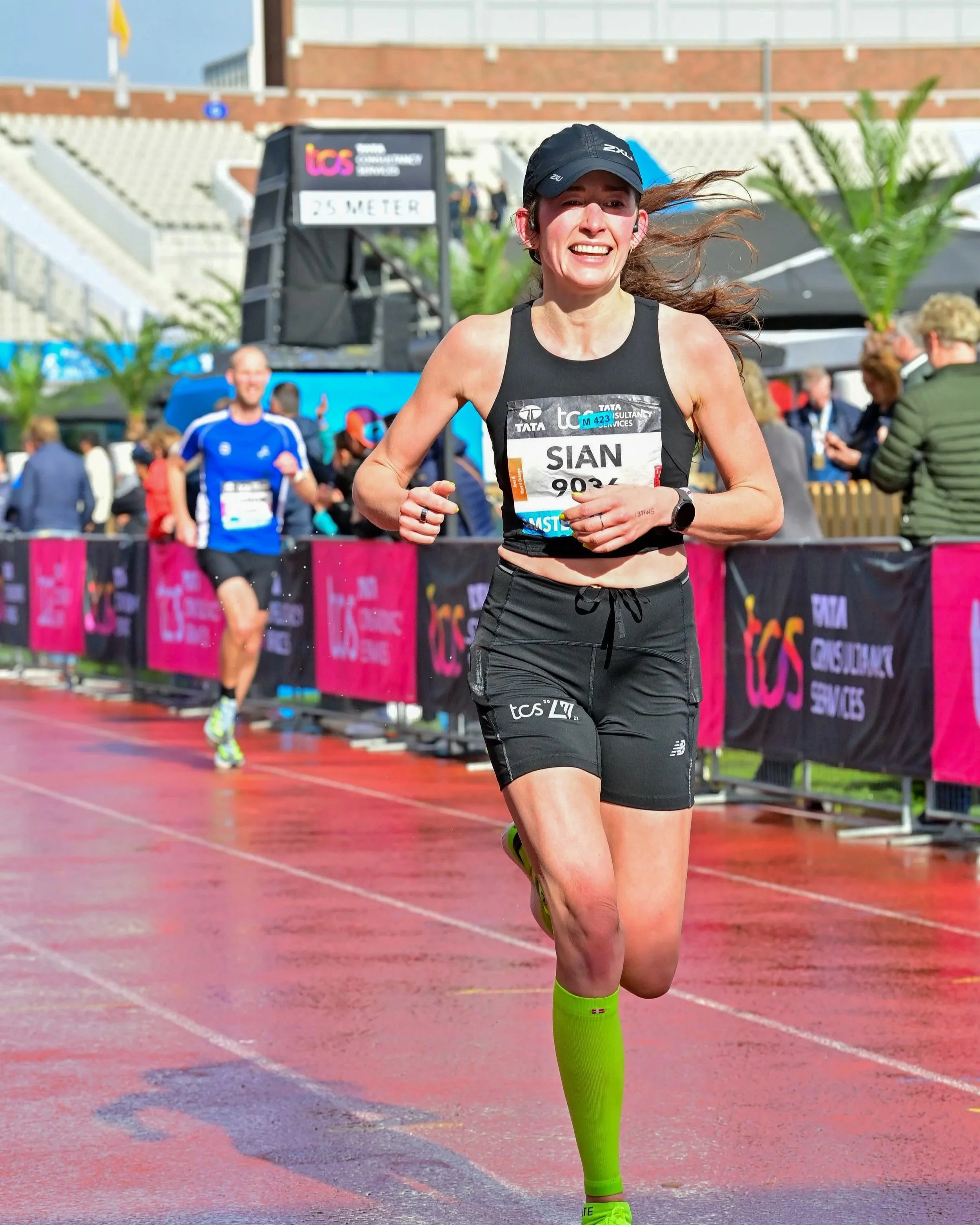 A female marathon runner in black sportswear and bright green compression socks running on a wet track during a race, smiling and nearing the finish line, with spectators and other runners in the background.