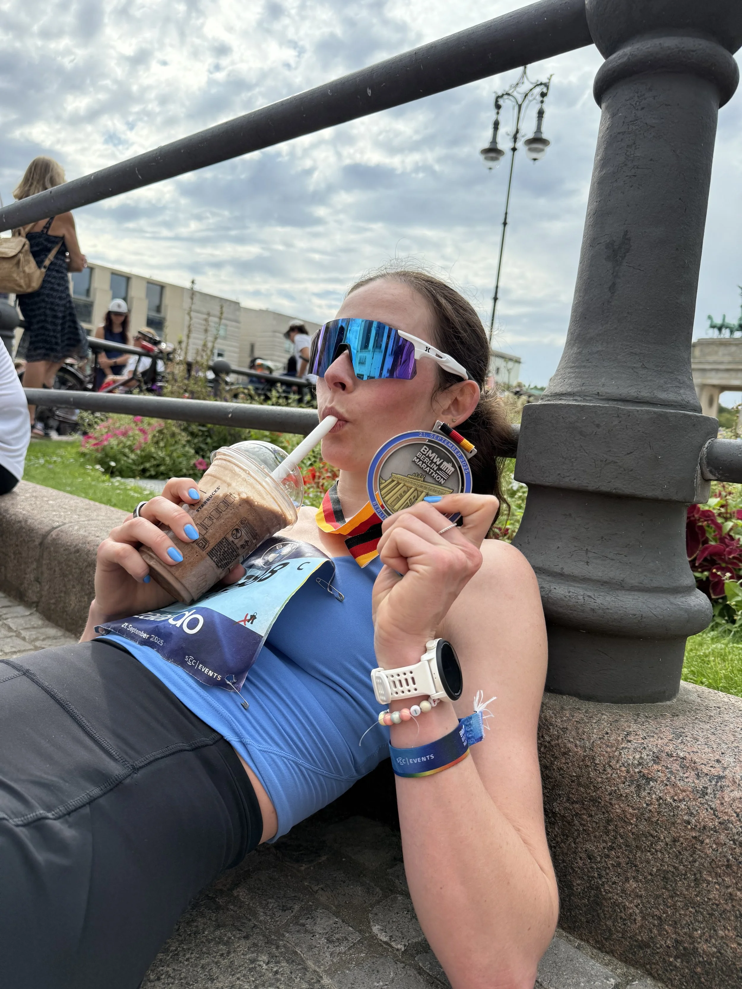 A woman lying on the ground, wearing sports attire and sunglasses, holding a medal in one hand and a chocolate smoothie in a cup with a straw in her other hand. She appears to be resting after a race, with a city street and other people in the background.
