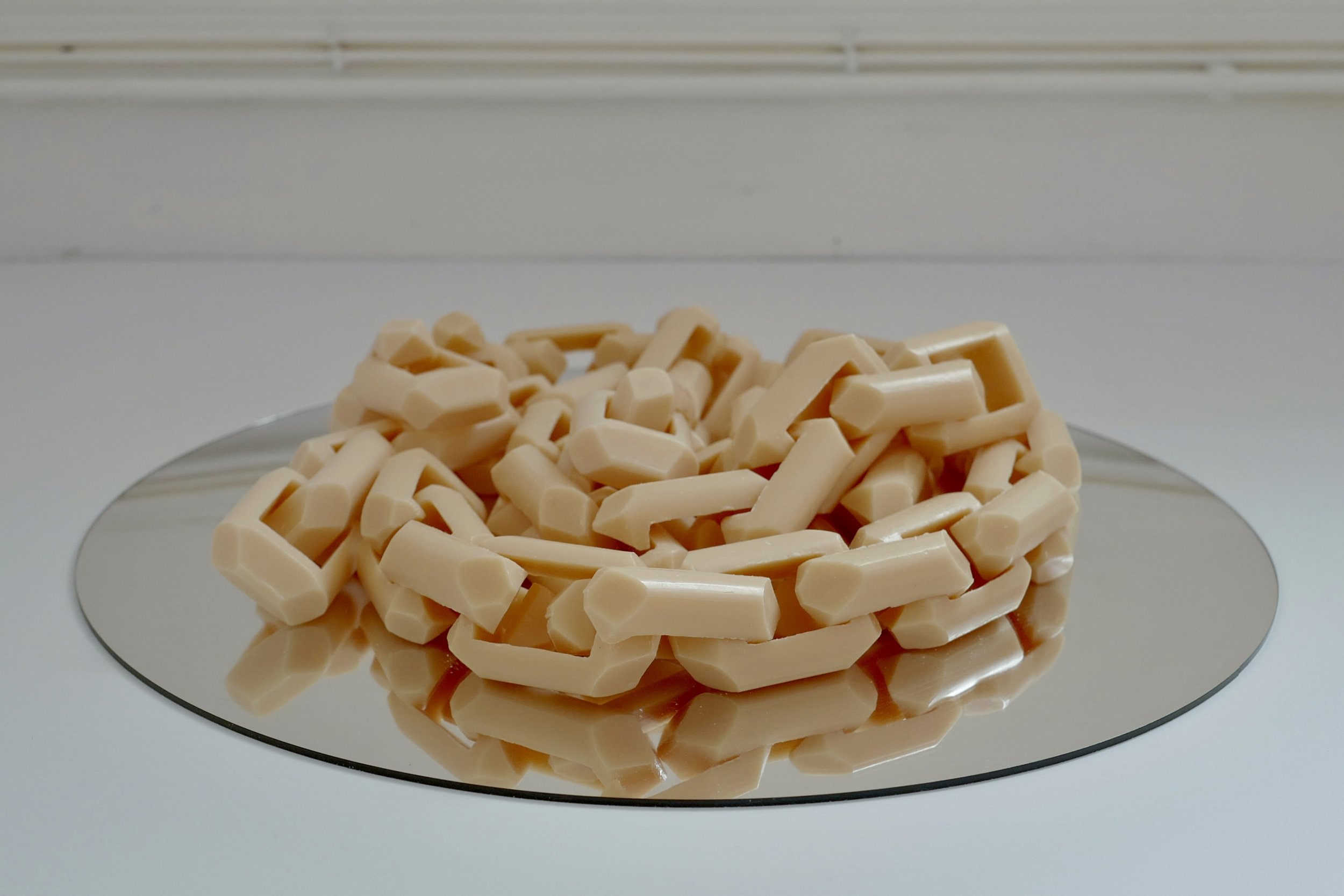 A large chain sits on top of a circular silver mirror. The links of the chain are made from individually carved bars of soap, connected to form a continuous chain. The sculpture is photographed on a plain white background in a gallery setting.