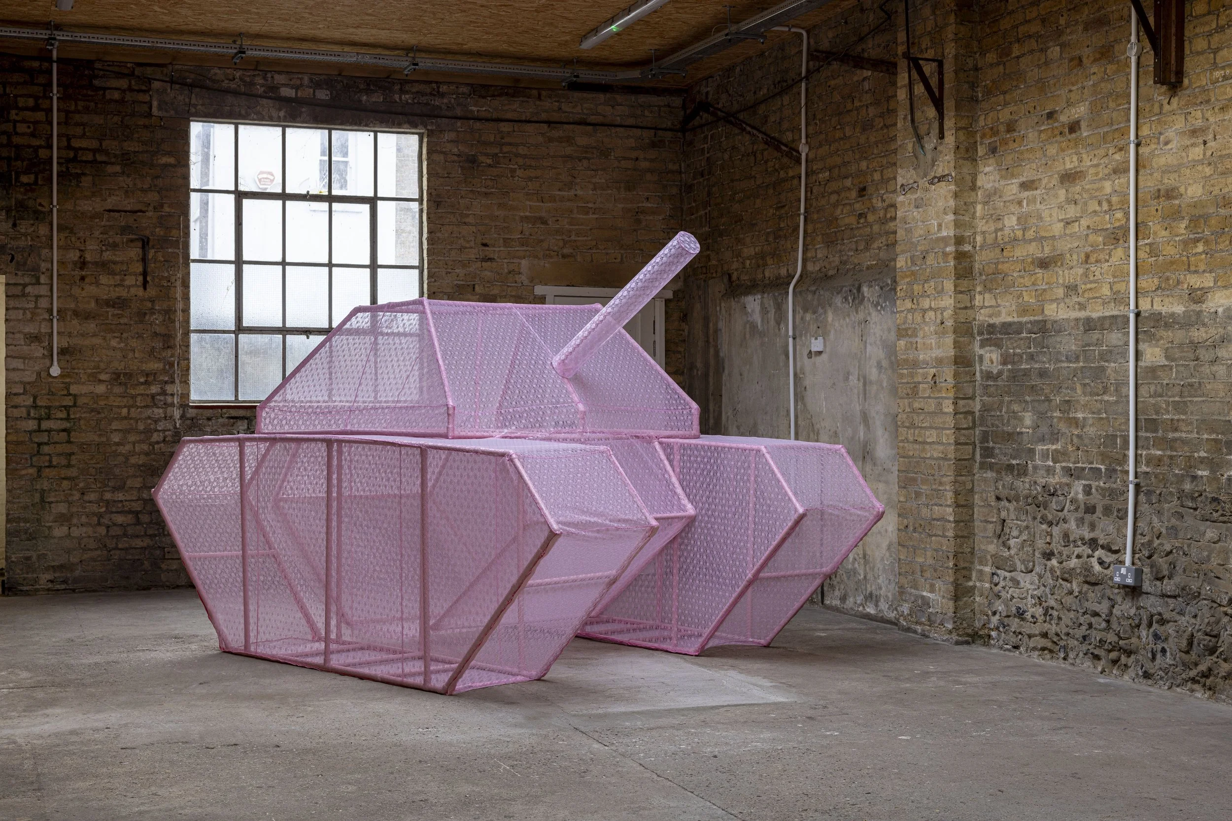 An image of a large-scale sculptural skeletal military tank covered in a pink lace fabric skin. The sculpture is photographed in an industrial warehouse setting with light visible passing through the ghost-like structure.