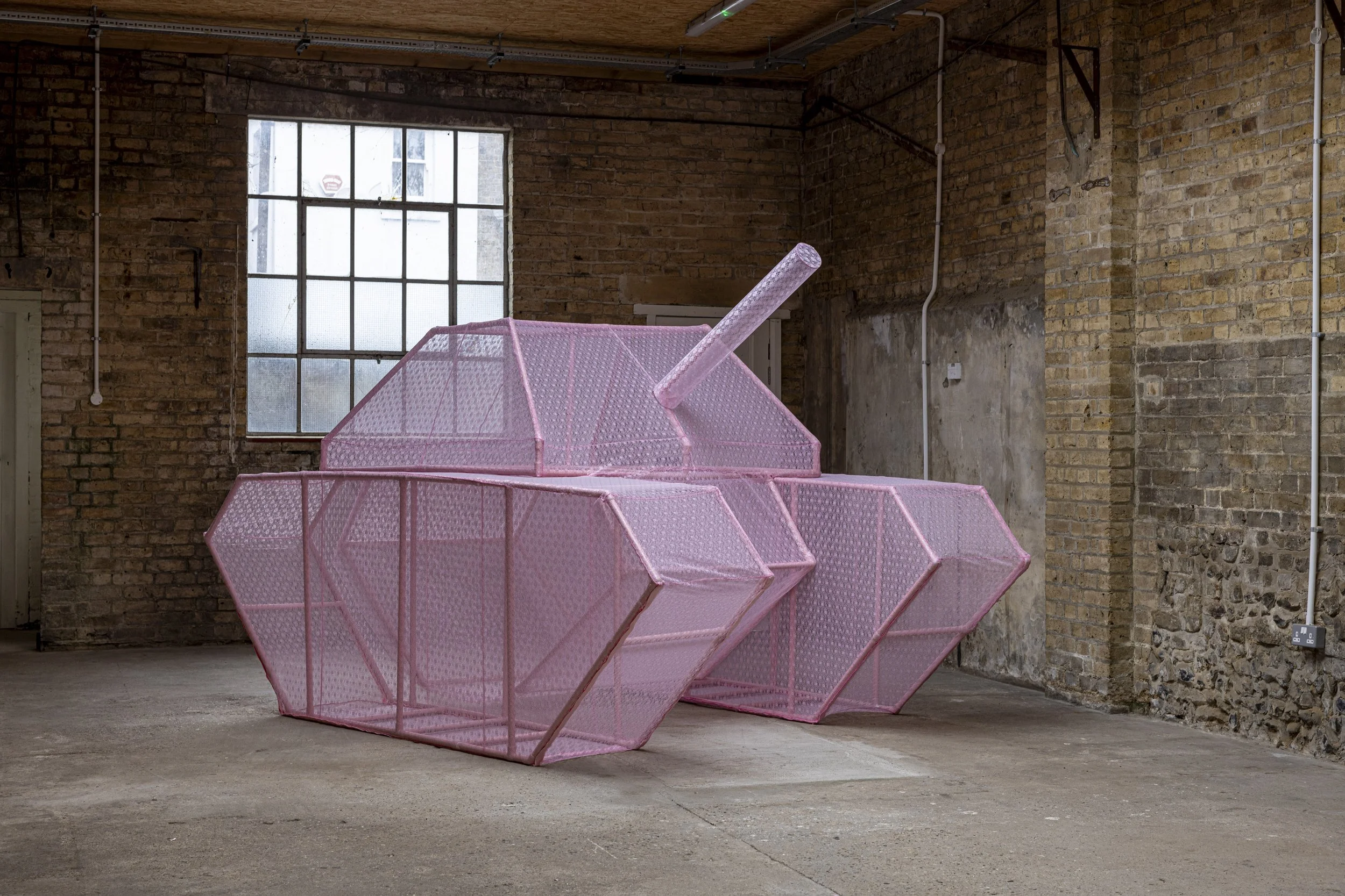 An image of a large-scale sculptural skeletal military tank covered in a pink lace fabric skin. The sculpture is photographed in an industrial warehouse setting with light visible passing through the ghost-like structure.