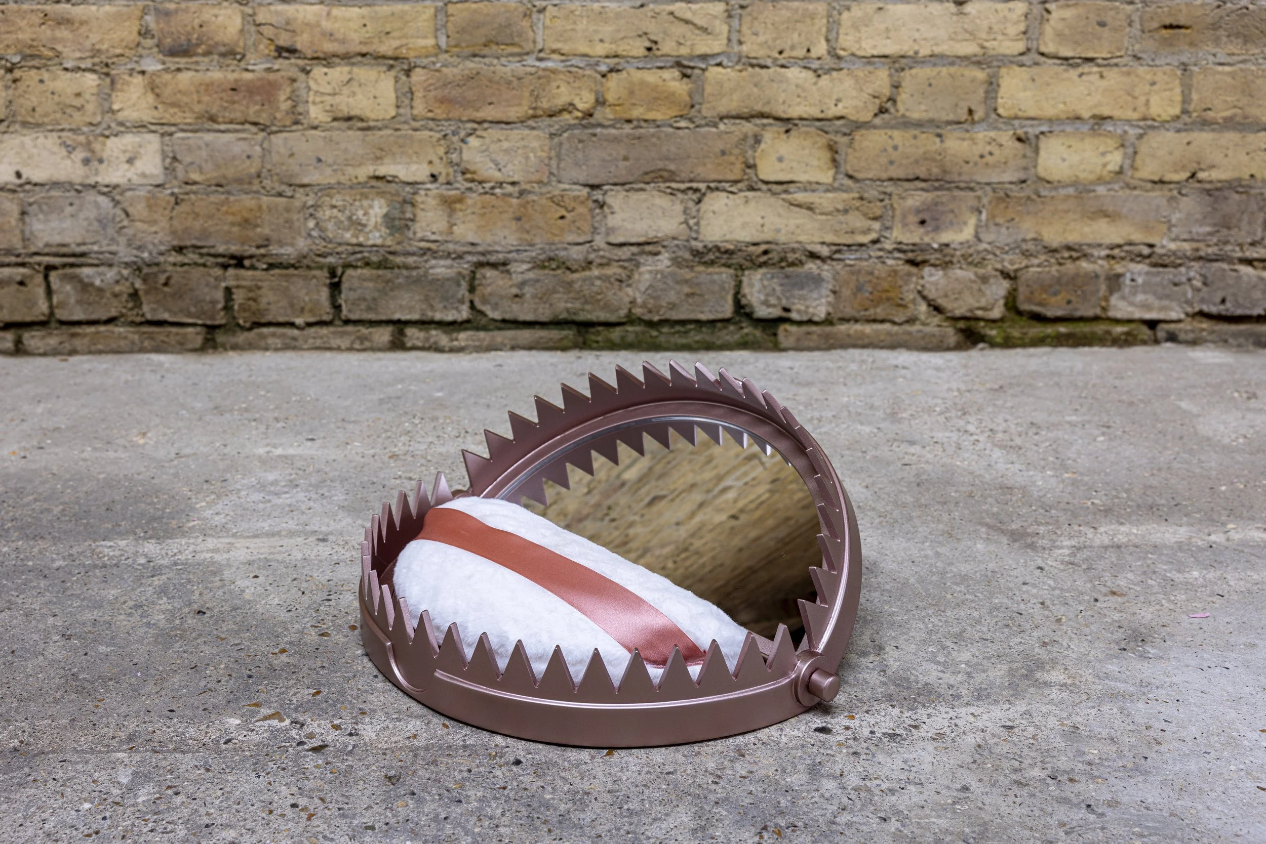 An image of a sculptural work which resembles a hybrid cosmetic powder compact with internal mirror, and a bear trap. It is metallic rose-gold in colour and contains a fluffy powder-puff inside. It is photographed on the ground in an industrial room.