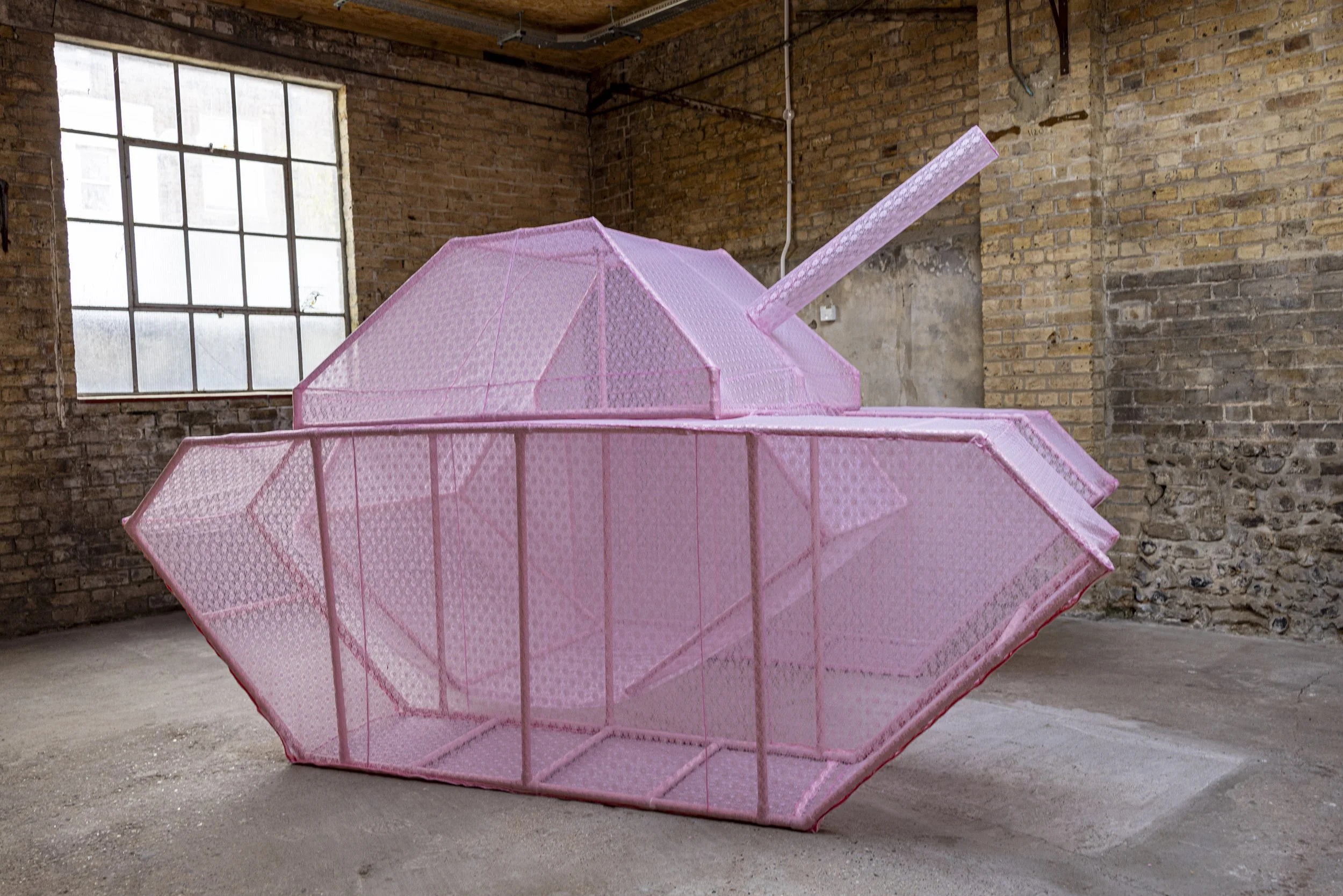 An image of a large-scale sculptural skeletal military tank covered in a pink lace fabric skin. The sculpture is photographed in an industrial warehouse setting with light visible passing through the ghost-like structure.