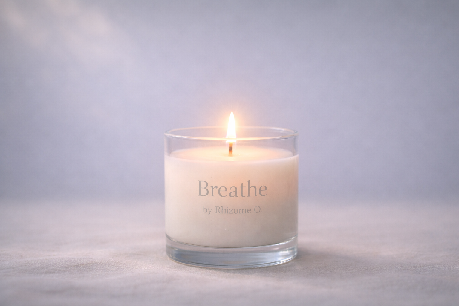 A lit white candle in a glass holder with the words 'Breathe by Rhizome O.' written on it.