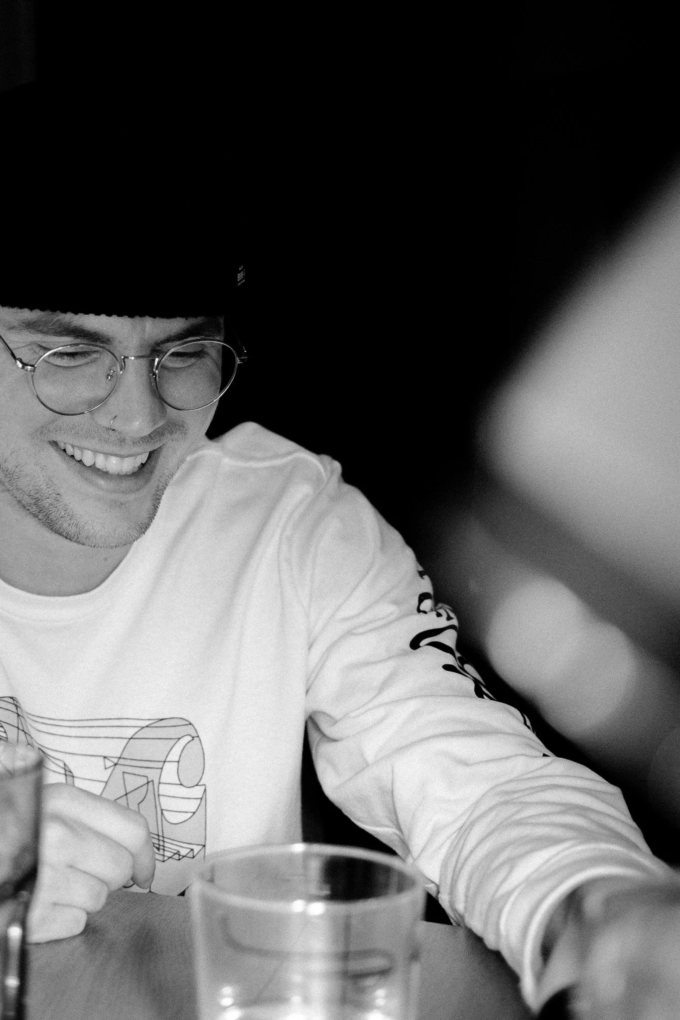 Un homme souriant portant des lunettes rondes, une casquette noire et un t-shirt blanc avec un dessin, assis à une table avec un verre dans la main, dans un environnement intérieur