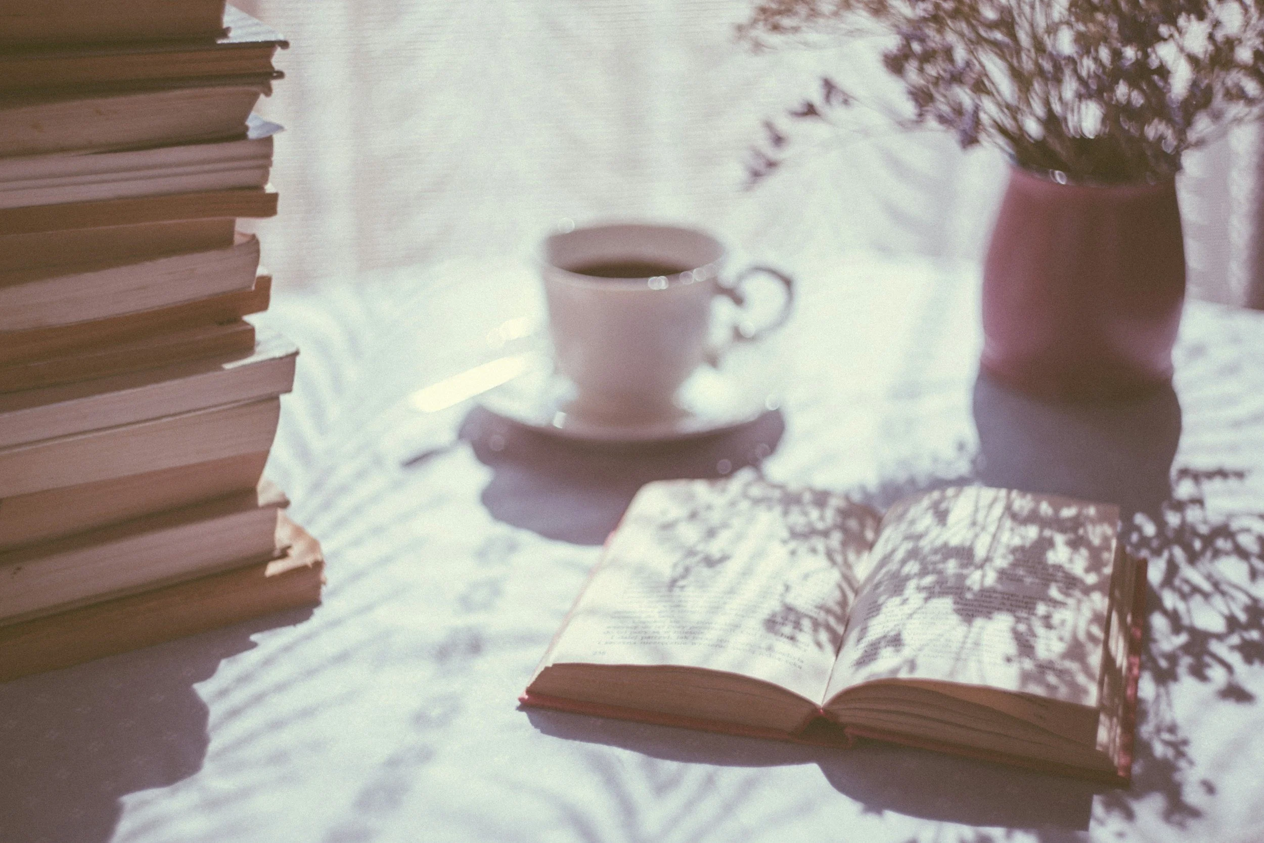 A table with a stack of books, an open book, a cup of coffee on a saucer, and a vase with flowers, all illuminated by sunlight.