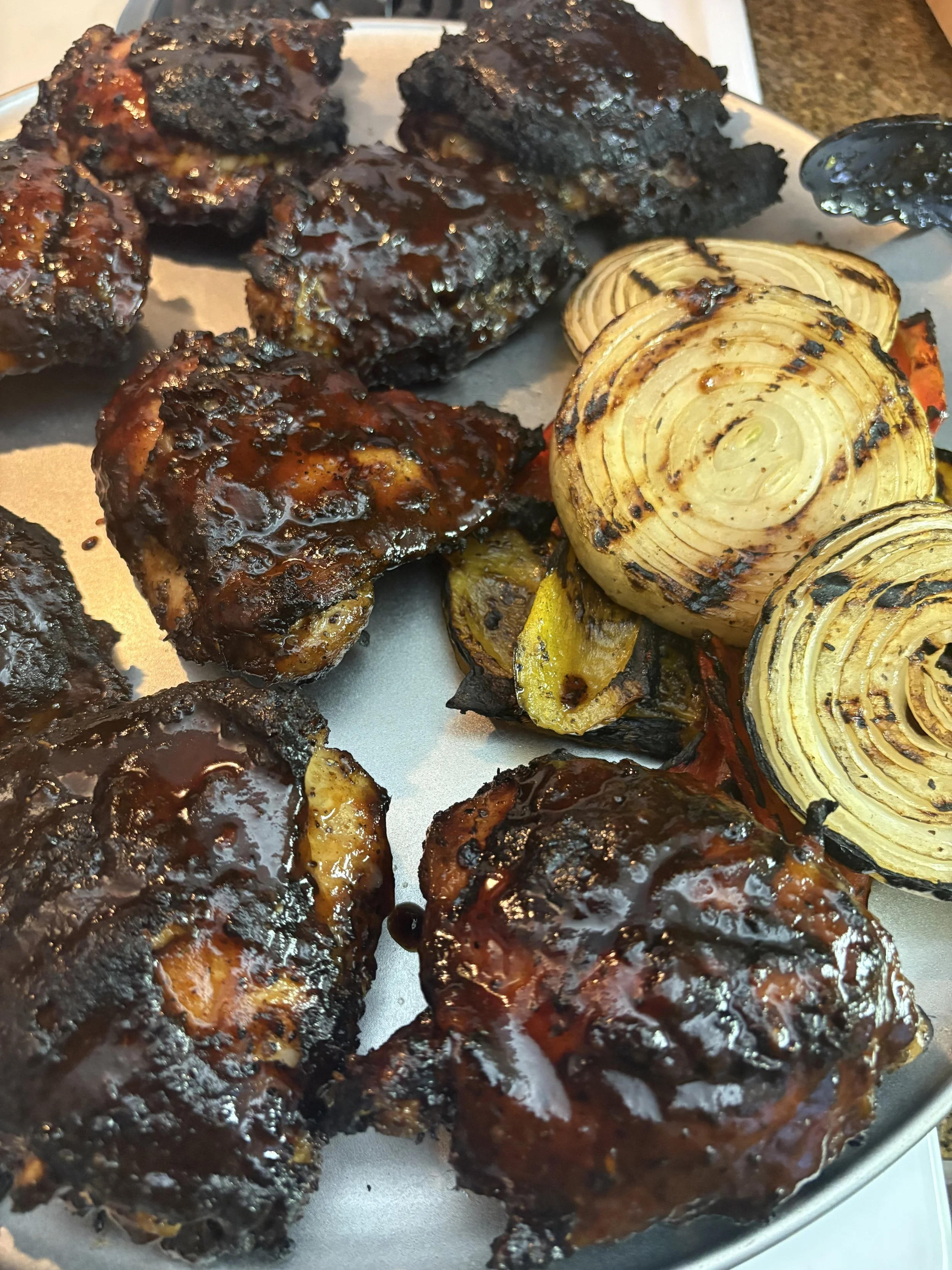 Barbecue chicken drumsticks with a dark, glossy sauce and charred edges, served on a tray with grilled onions and peppers.