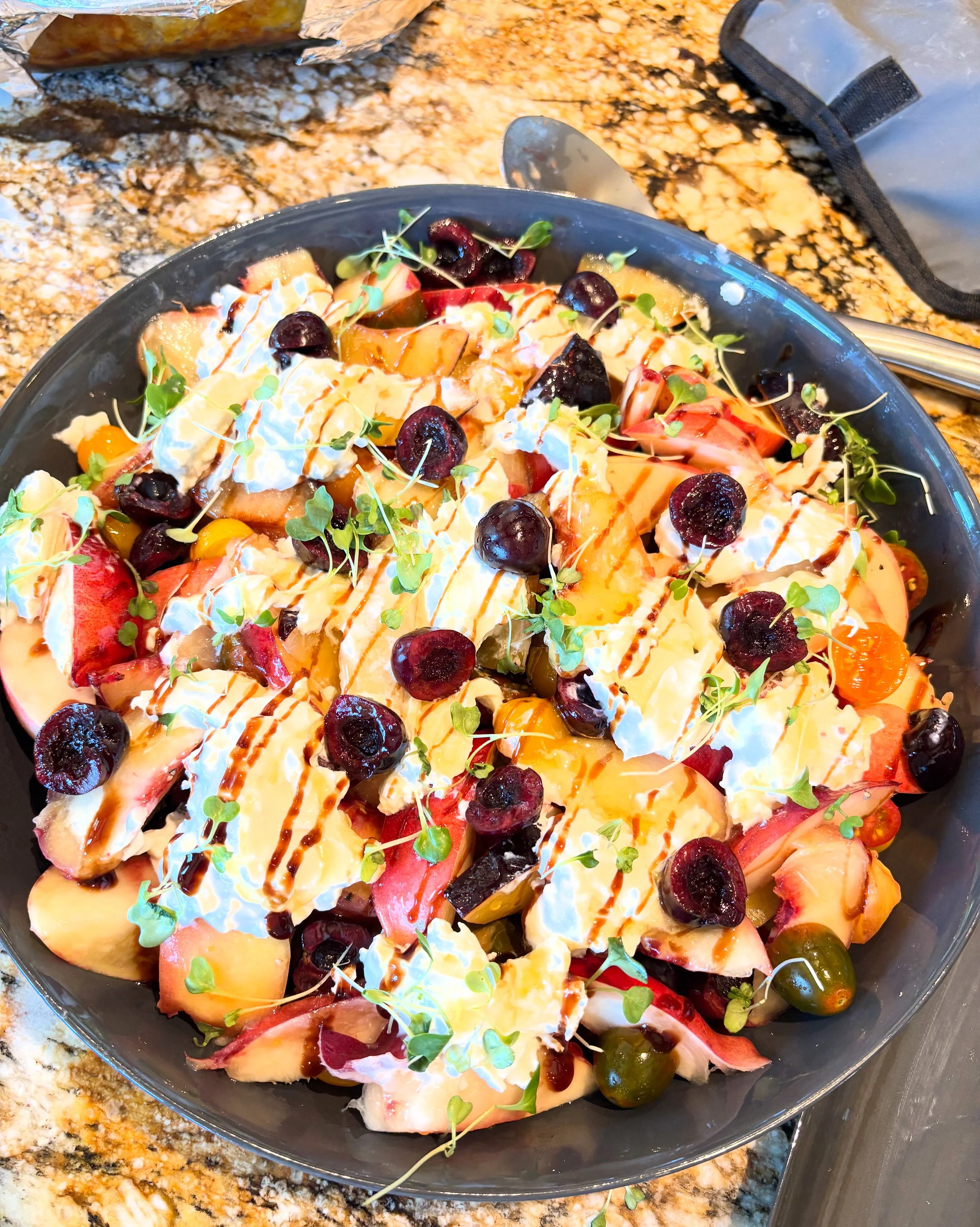 A fresh fruit salad with blueberries, peaches, strawberries, and grapes topped with microgreens and drizzled with balsamic glaze in a black bowl on a granite countertop.