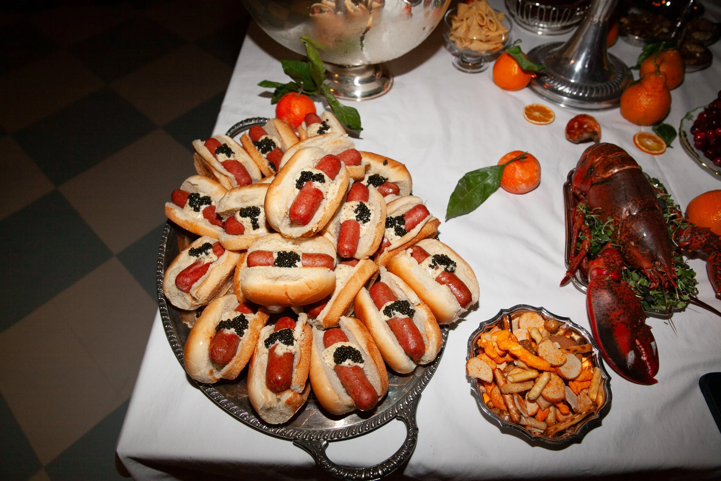 A silver tray with mini hot dogs in buns topped with black caviar, set on a white tablecloth with oranges and a lobster nearby.