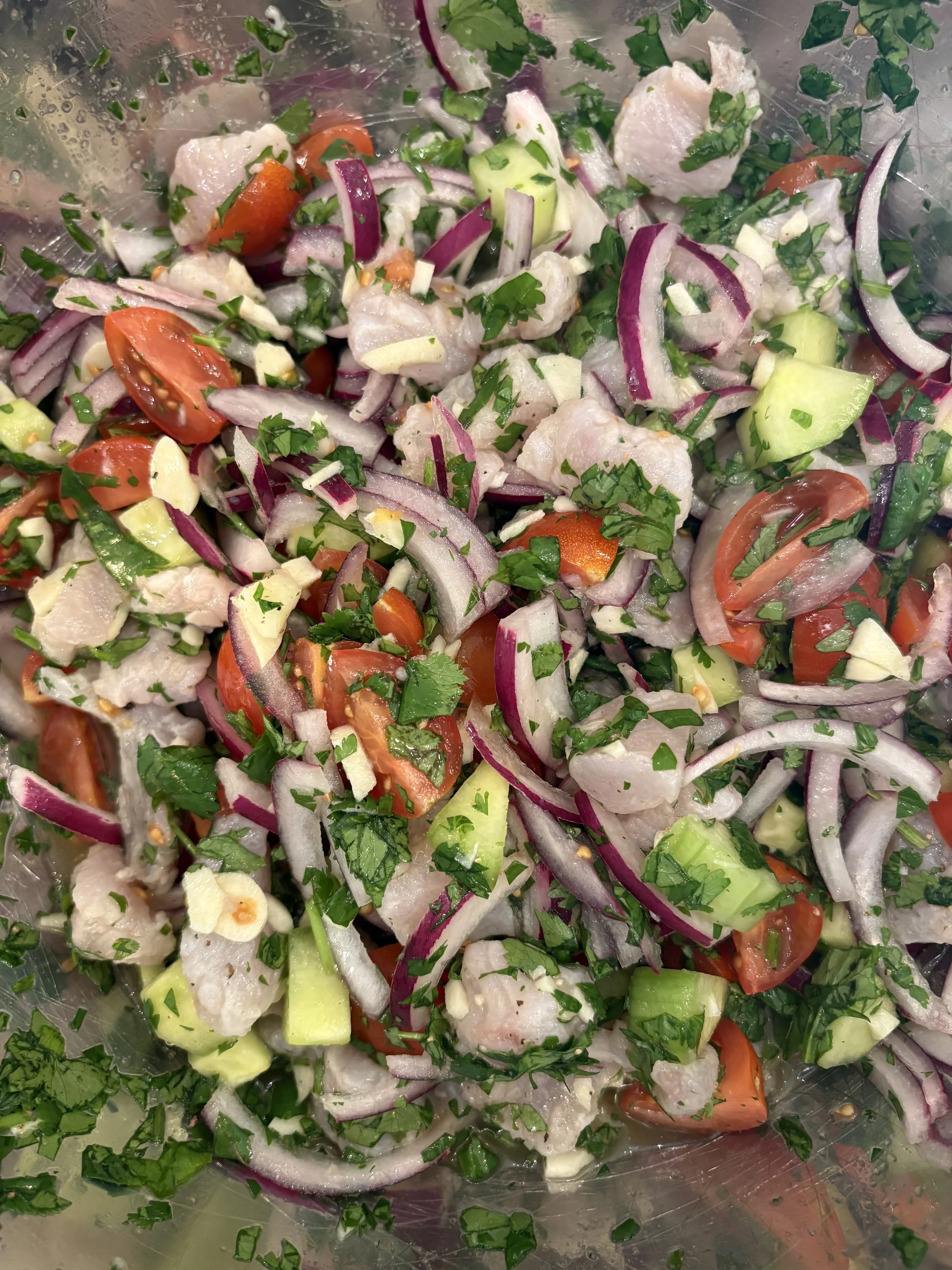 A bowl of chopped vegetables and raw fish, including red onions, cherry tomatoes, cucumbers, garlic, parsley, and fish fillets.