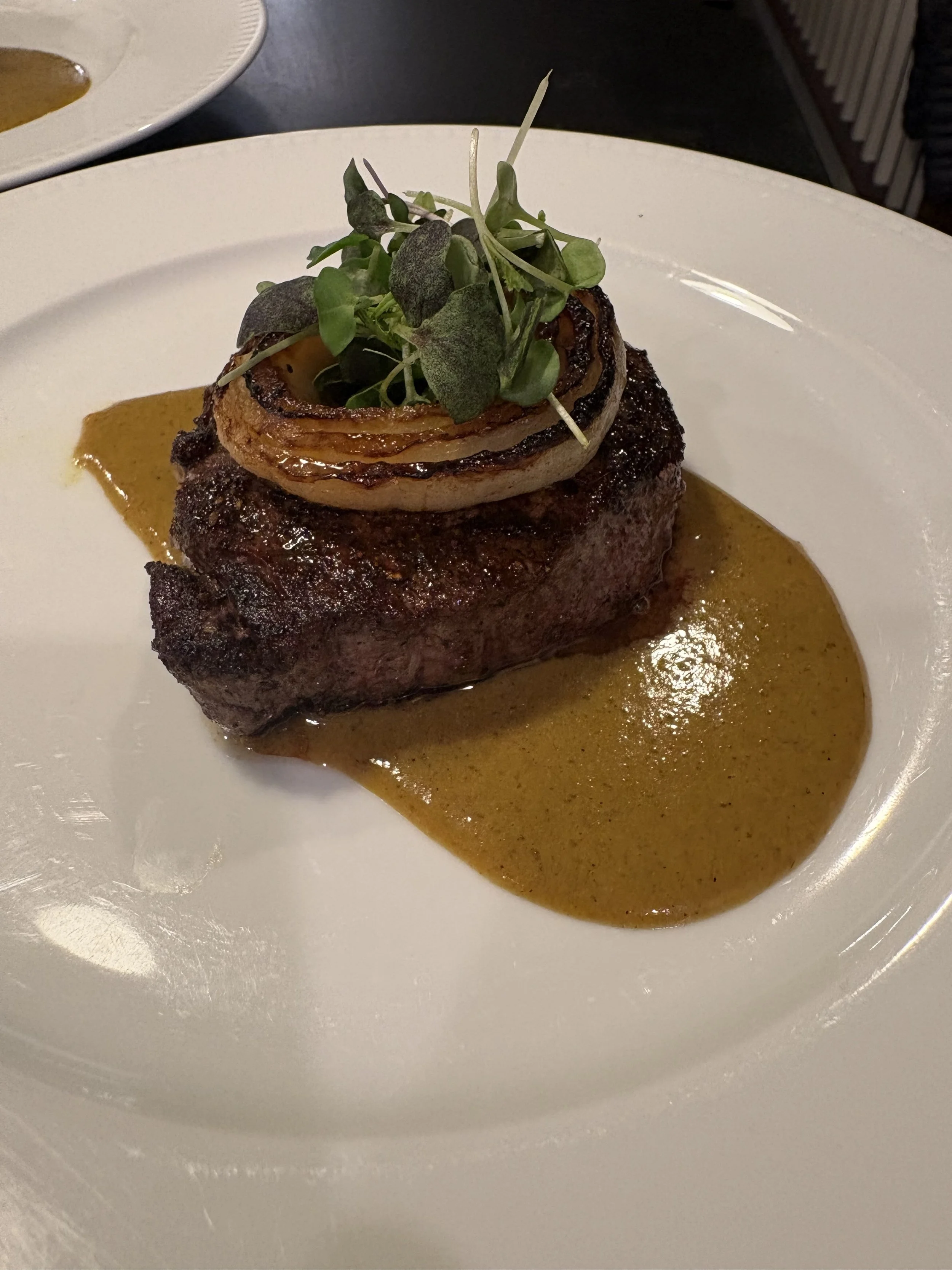 A steak topped with grilled onion and microgreens, served with a brown sauce on a white plate.