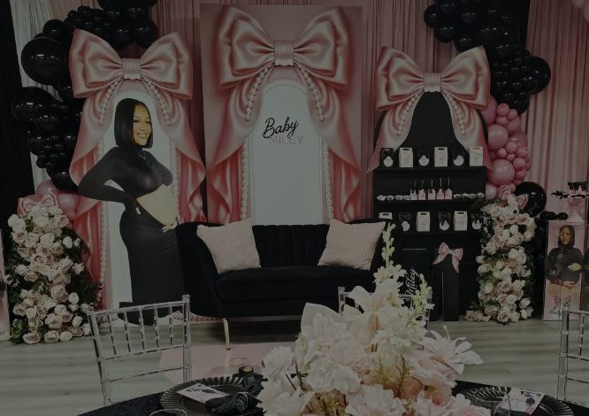Decorative event setup with pink and black balloon arches, large pink bows, and floral arrangements. A framed photo of a woman and the words 'Baby Faith' are in the background, indicating a baby shower theme.