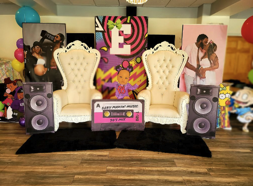 Decorative backdrop with two white ornate chairs, large speakers, and cartoon character cutouts for a child's birthday party, with colorful balloons and a poster with a bear and the letter 'E' behind.