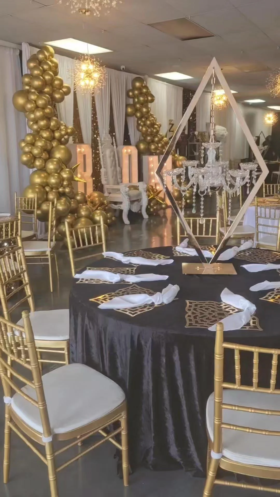 Decorated event space with black and gold theme including gold balloons, gold chairs, a black tablecloth with gold accents, a crystal chandelier, and a white backdrop with white curtains.
