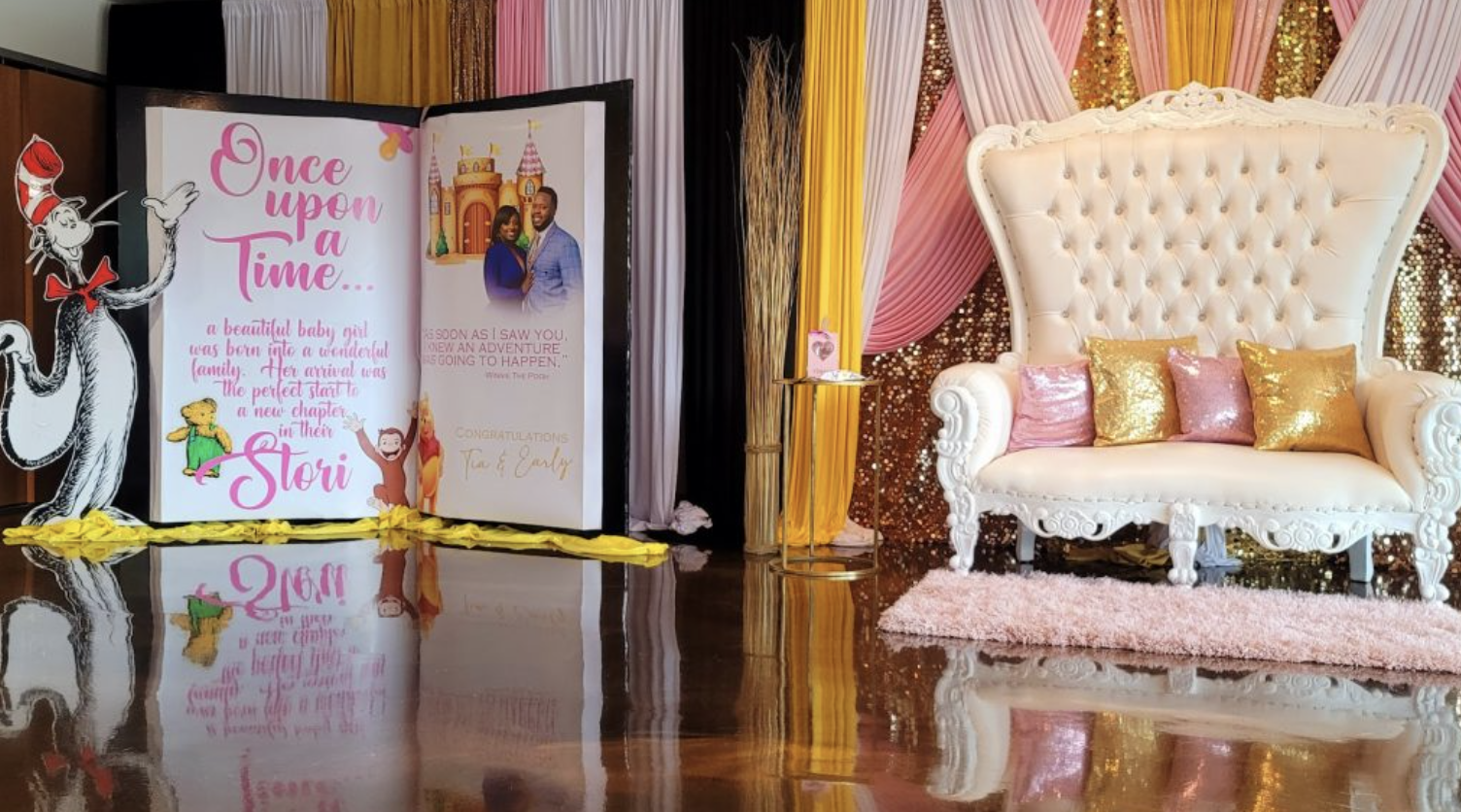 Decorative setup for a baby shower with a book display, a white ornate sofa with pink and gold pillows, pink and gold draped fabric backdrop, and themed decorations including a cartoon mouse with a party hat.