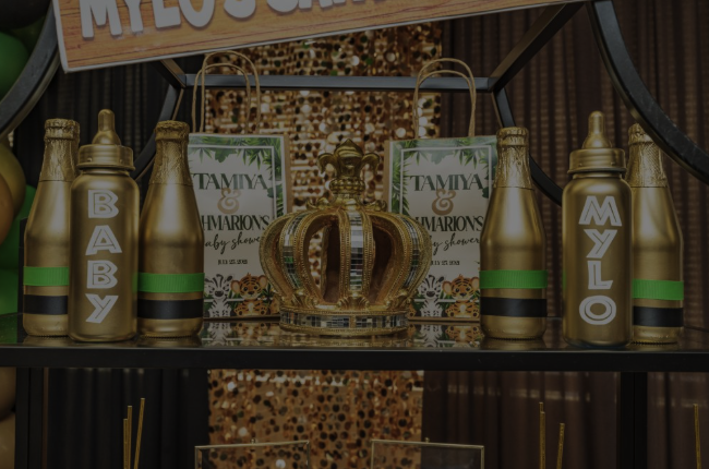 Gold-colored baby bottles with green stripes spelling B A B Y M Y L O, a gold crown, and decorative signs on a display table.