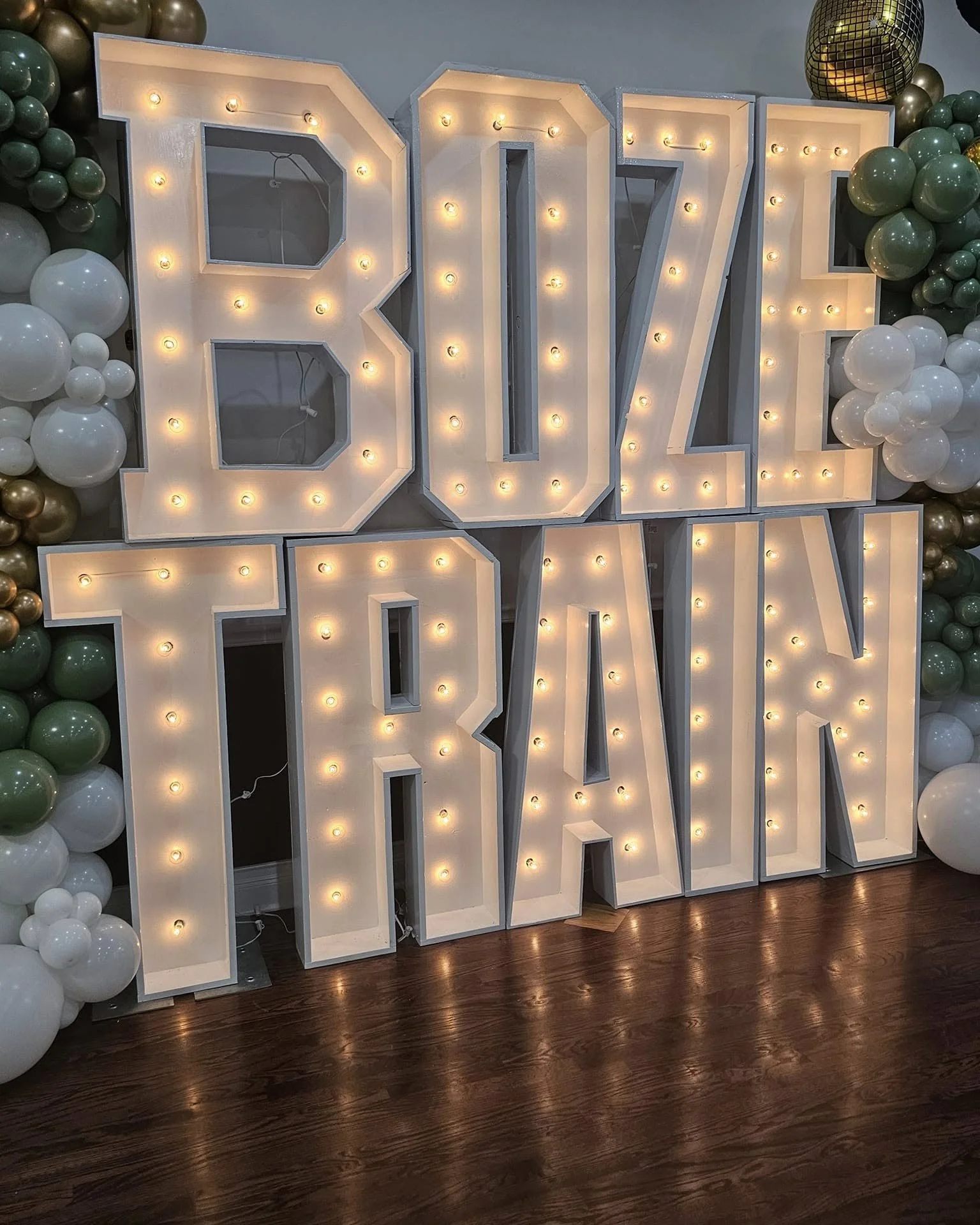 Large illuminated sign spelling out 'ONE YEAR' with a backdrop of green, white, and gold balloons.