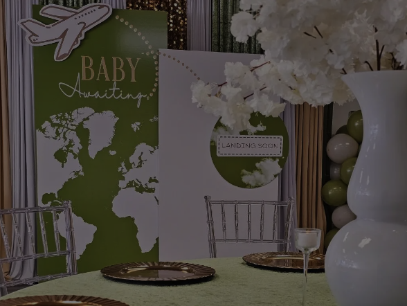 Decorative baby shower table setup with green and white theme, including a large white vase with white flowers, a green and white sign with clouds and the text "Landing Soon," and a green sign with a world map and the text "Baby Awaiting."