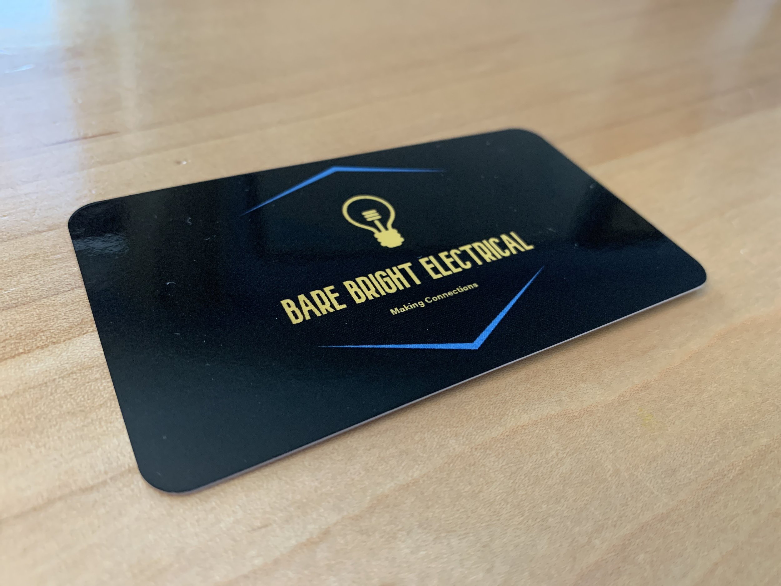 Black business card for Bare Bright Electrical on a wooden surface, with a yellow lightbulb logo and blue accents.