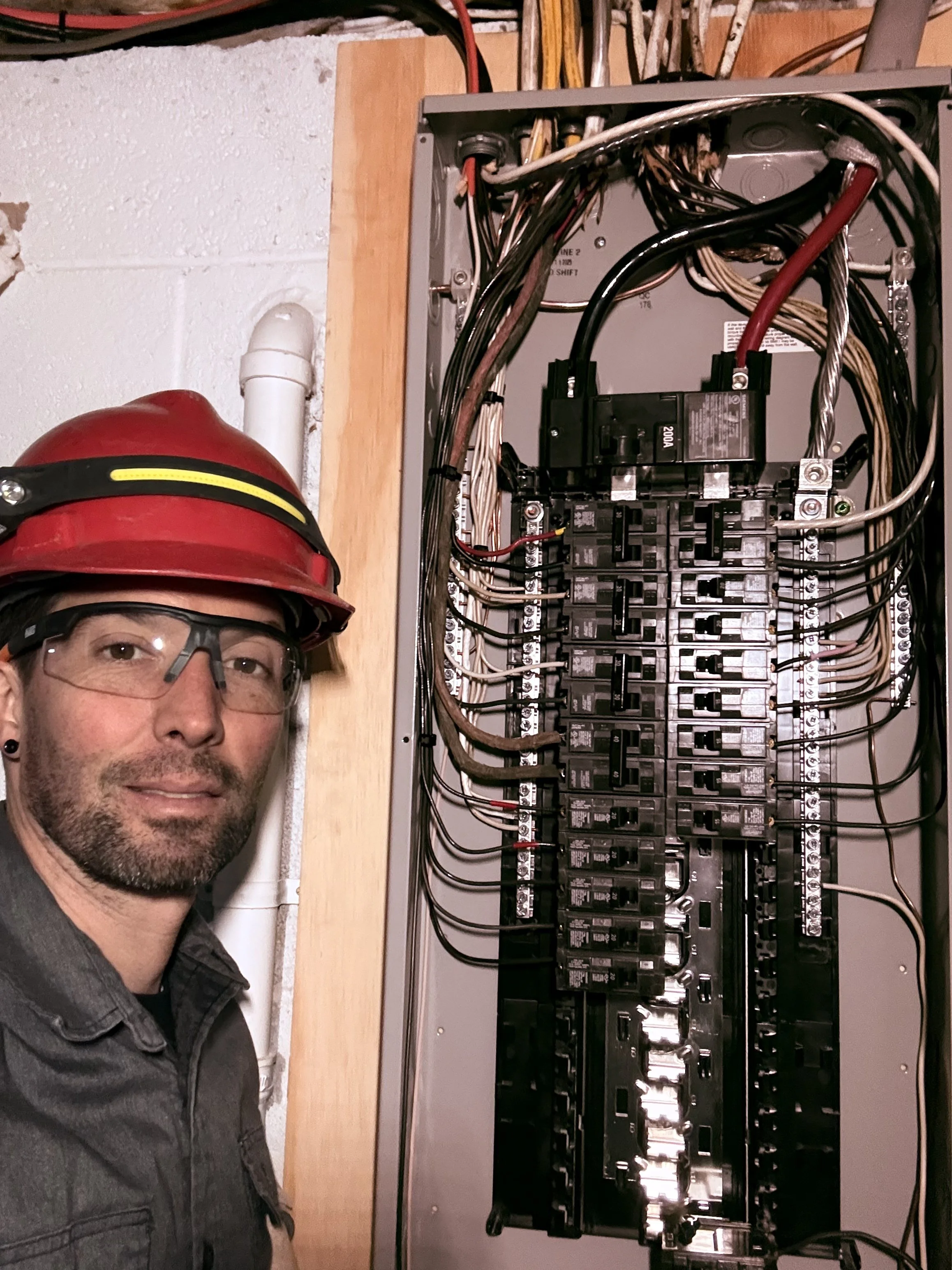 A man wearing a red helmet, safety glasses, and a gray shirt standing next to an open electrical panel with circuit breakers and wiring.