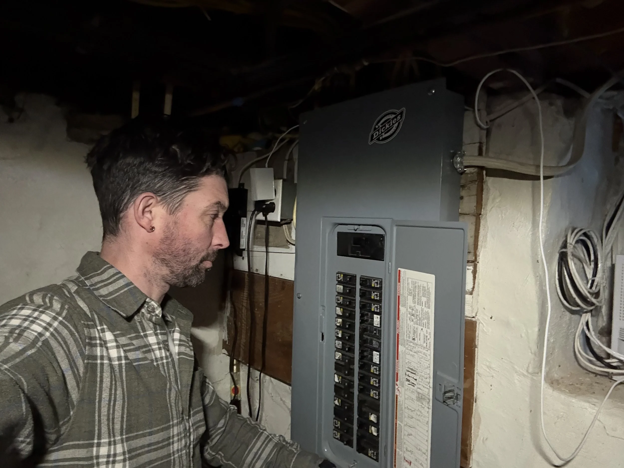 A man inspecting an electrical circuit breaker panel in a basement or utility room.